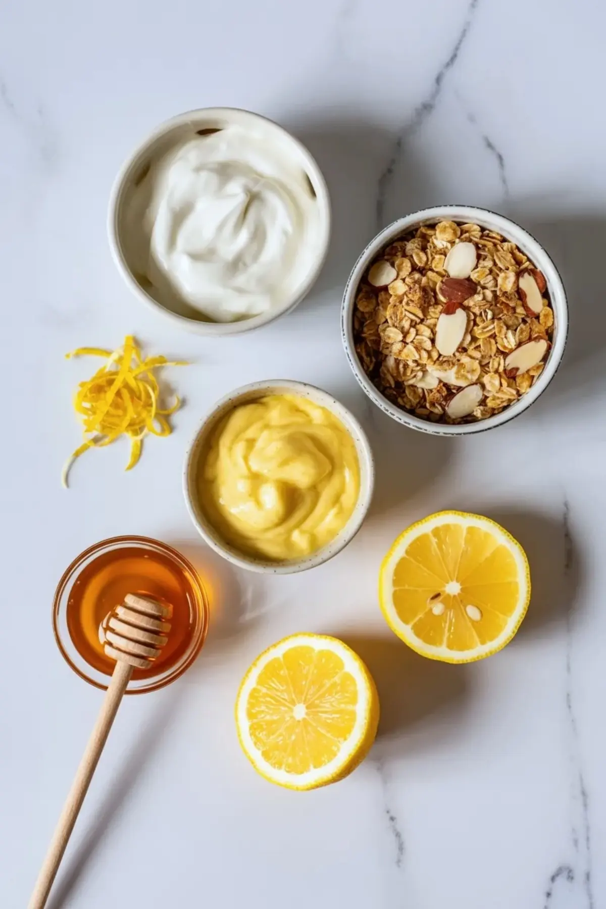 Ingredients for lemon yogurt parfait with granola arranged in small bowls with lemon curd, Greek yogurt, honey, granola, lemon zest, and lemon halves.