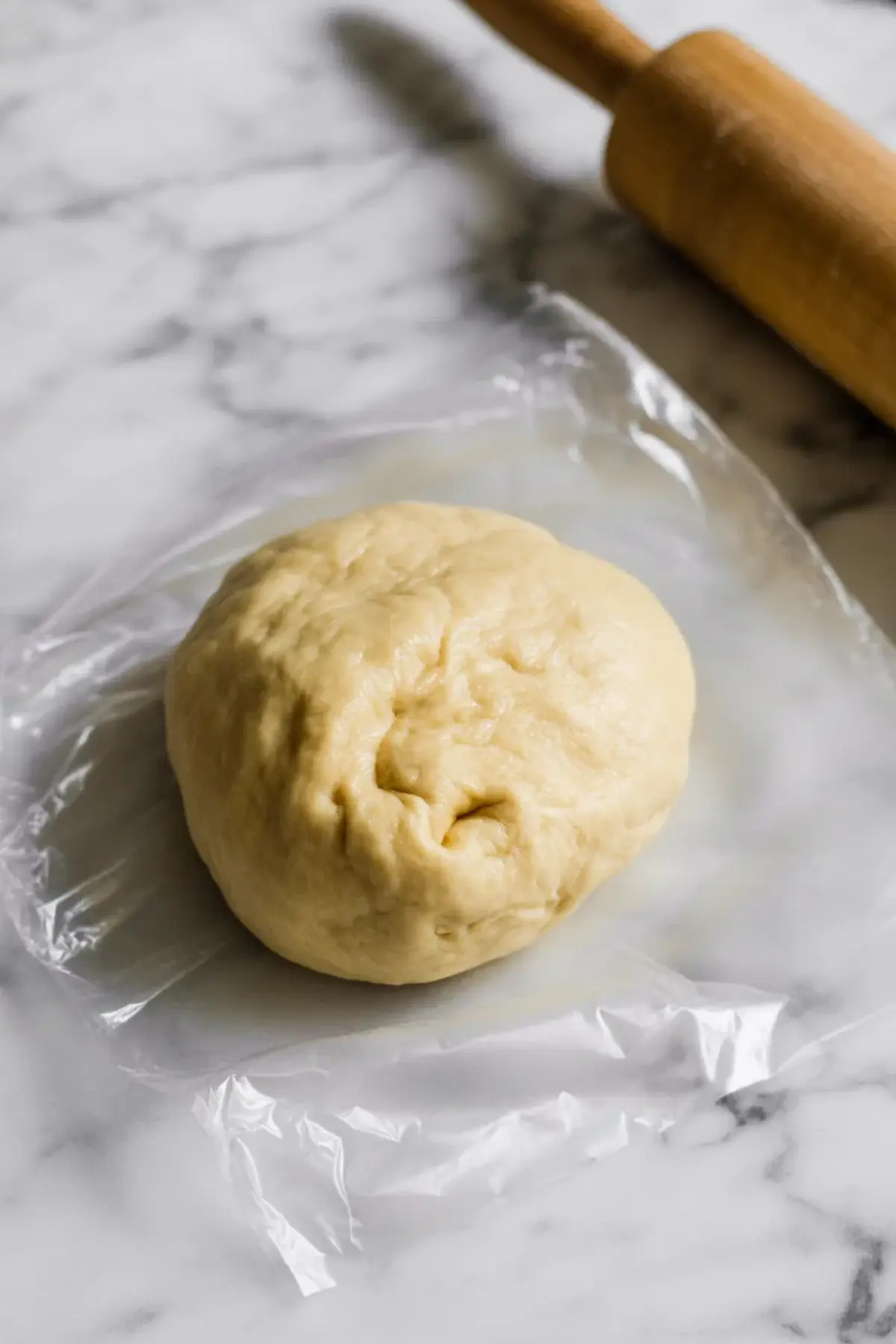Ball of homemade tart dough rests on plastic wrap on a marble countertop with a wooden rolling pin in the background, showing soft pastry texture ready for chilling, pie crust preparation, and step by step mango tart crust recipe.