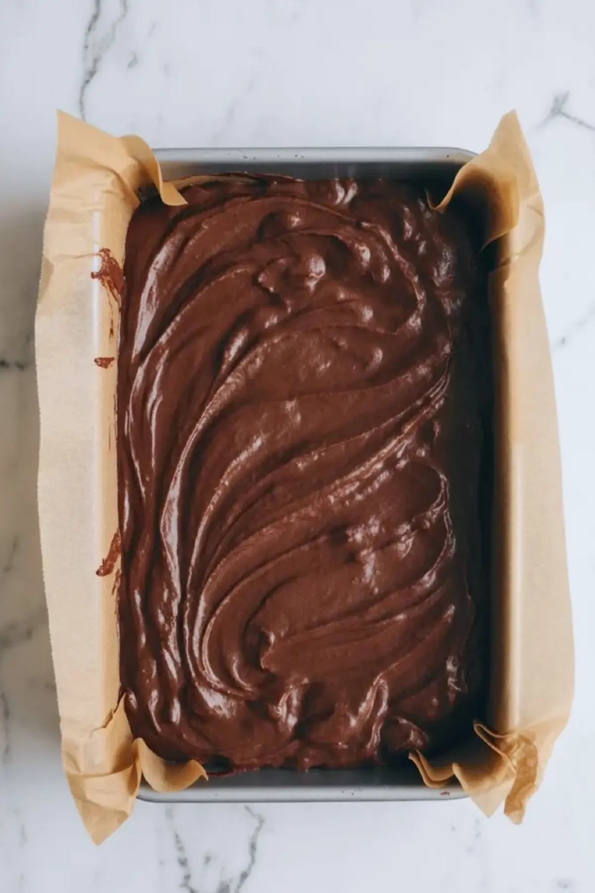 Chocolate brownie batter sits spread in a parchment-lined baking pan, showing the thick glossy base before the marshmallow brownies bake.