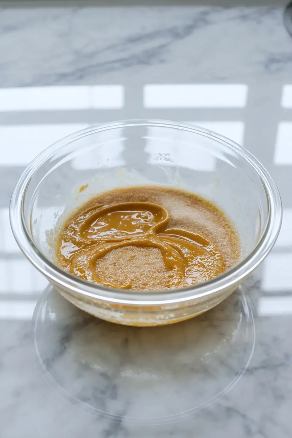 Melted butter and sugar sit mixed in a glass bowl on a marble surface, showing one of the early brownie base steps for marshmallow brownies.
