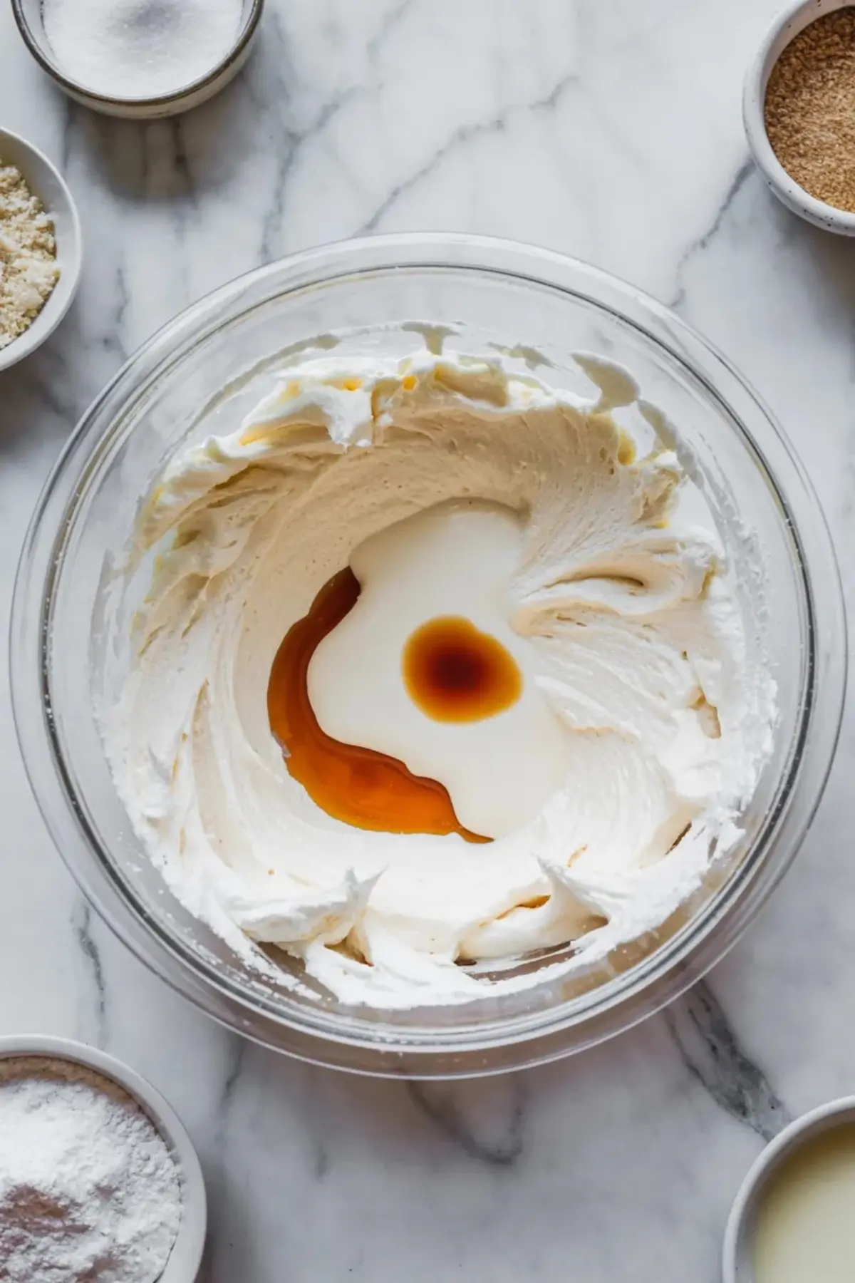 Glass mixing bowl holds whipped buttercream with vanilla extract and cream in the center on a marble countertop with small bowls of sugar and baking ingredients around the bowl, showing marshmallow buttercream frosting preparation.
