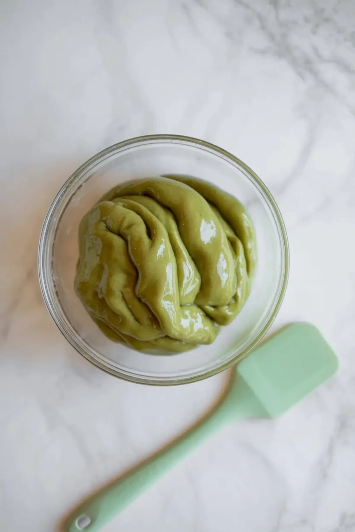 Glass bowl holds glossy green matcha mochi dough on a white marble surface with a mint silicone spatula beside the bowl, showing smooth sticky texture for homemade matcha mochi dessert preparation.
