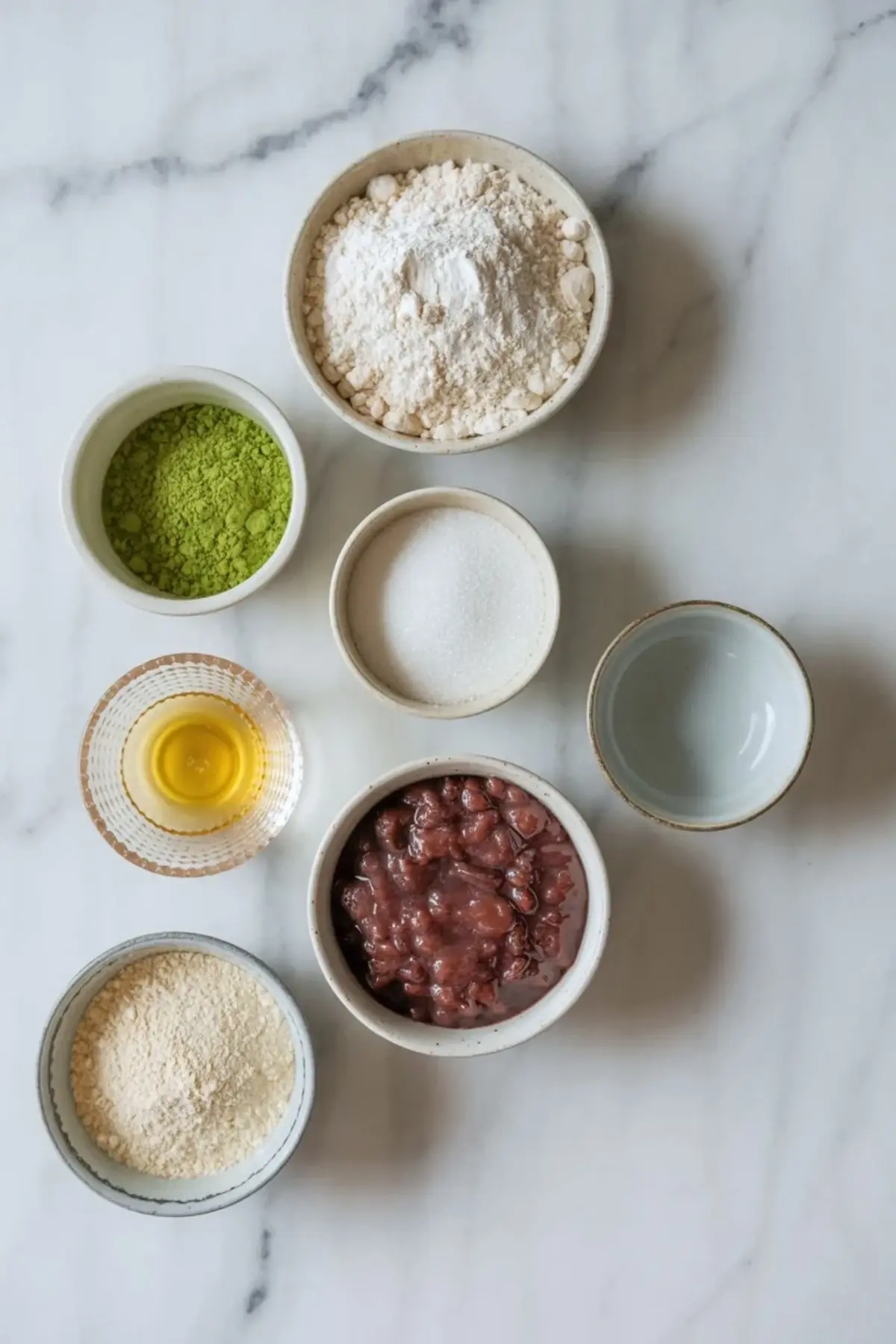 Small bowls display matcha powder, flour, sugar, oil, water, glutinous rice flour, and sweet red bean paste arranged on a marble surface as measured ingredients for matcha red bean mochi.
