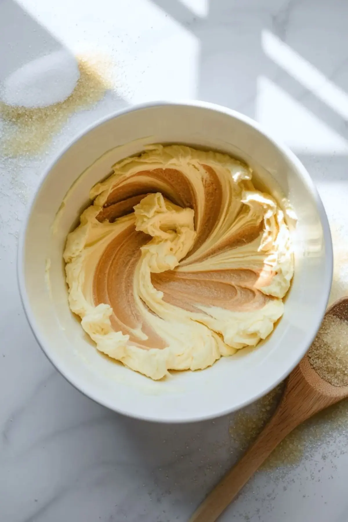 Bowl shows creamed butter and sugar mixture with smooth swirls, placed beside granulated sugar and a wooden spoon on a bright marble countertop.
