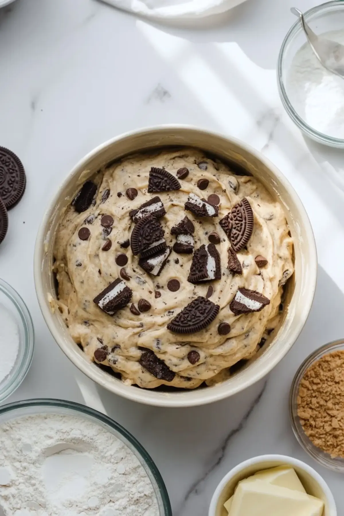 Bowl holds thick Oreo cookie dough with crushed Oreo pieces and chocolate chips on top, surrounded by baking ingredients like flour, butter, and sugar on a white marble surface.
