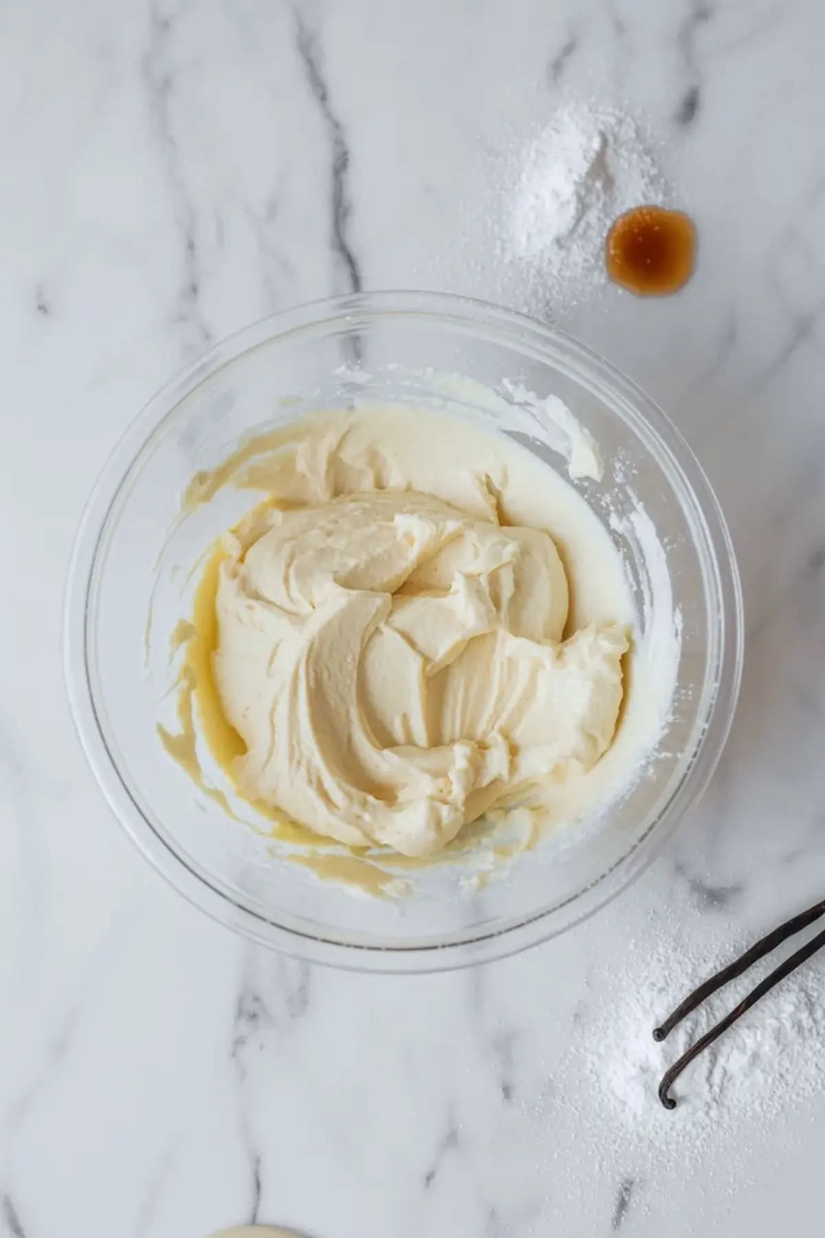 Cream cheese base for Oreo mousse in a clear bowl with vanilla beans and powdered sugar on white marble before the whipped cream is folded in.