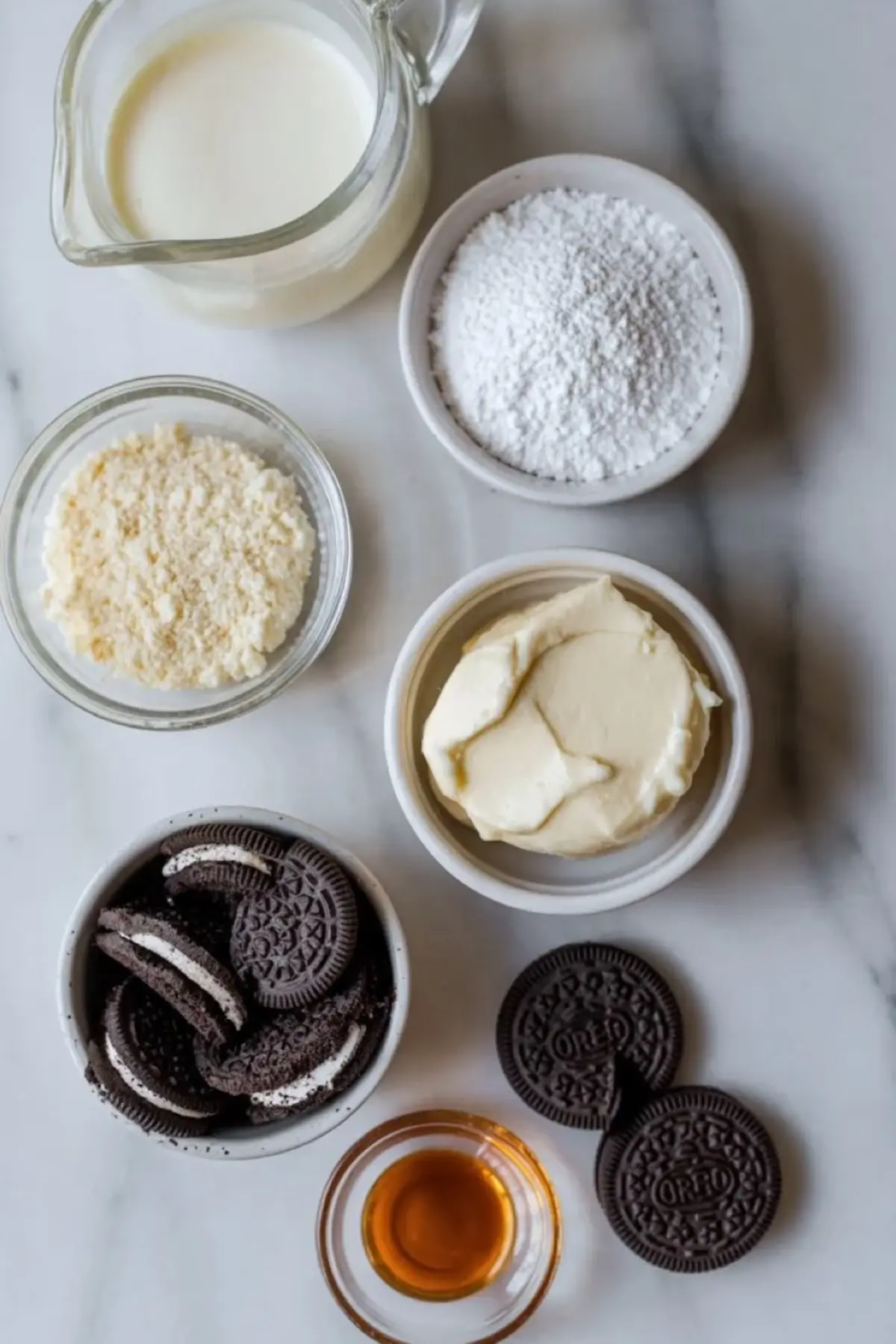 Ingredients for Oreo mousse arranged with heavy cream, powdered sugar, cream cheese, Oreo cookies, and vanilla on white marble.