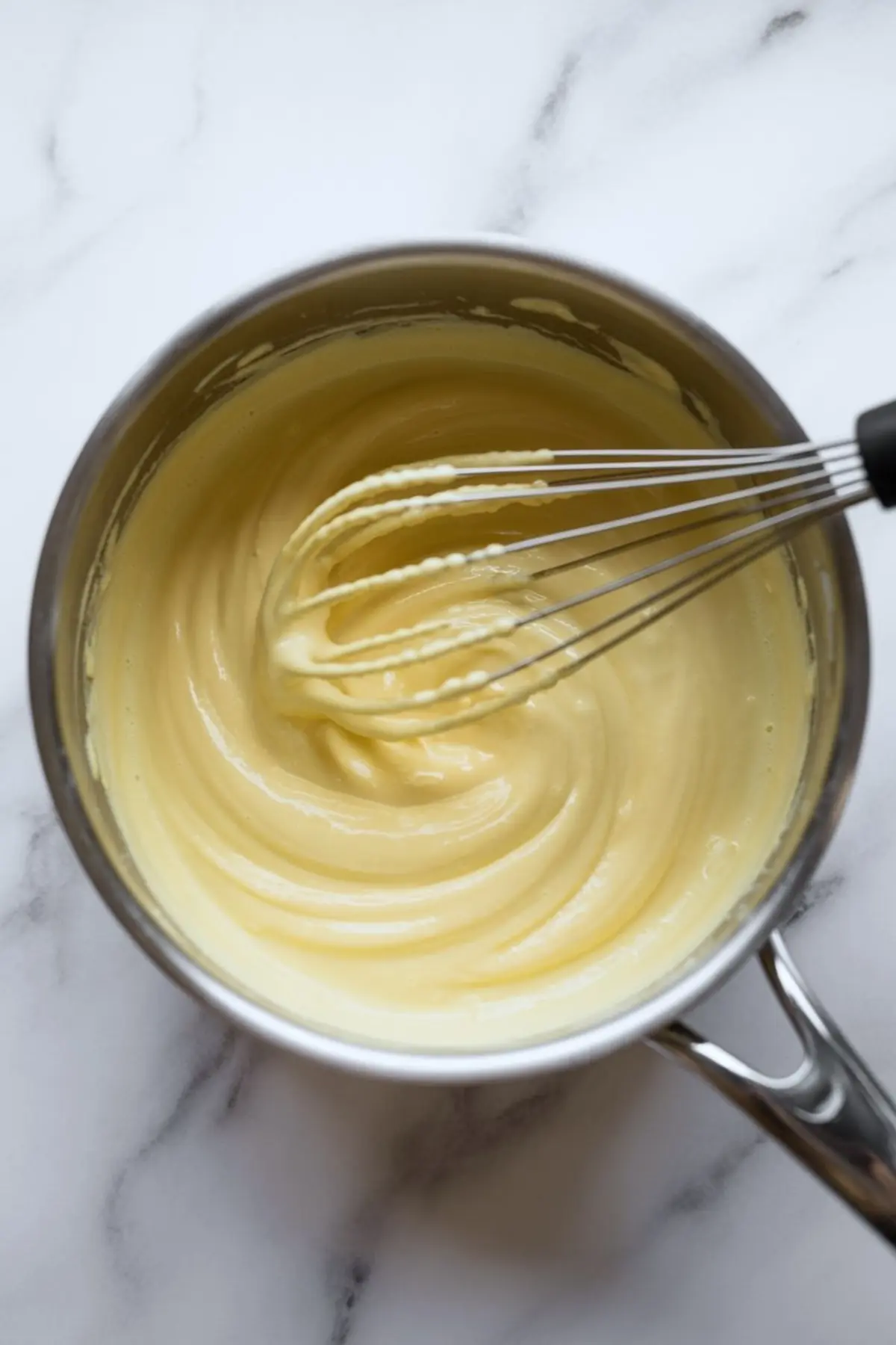 Whisk mixes smooth vanilla custard in stainless steel saucepan creating thick glossy pastry cream texture for homemade dessert filling.
