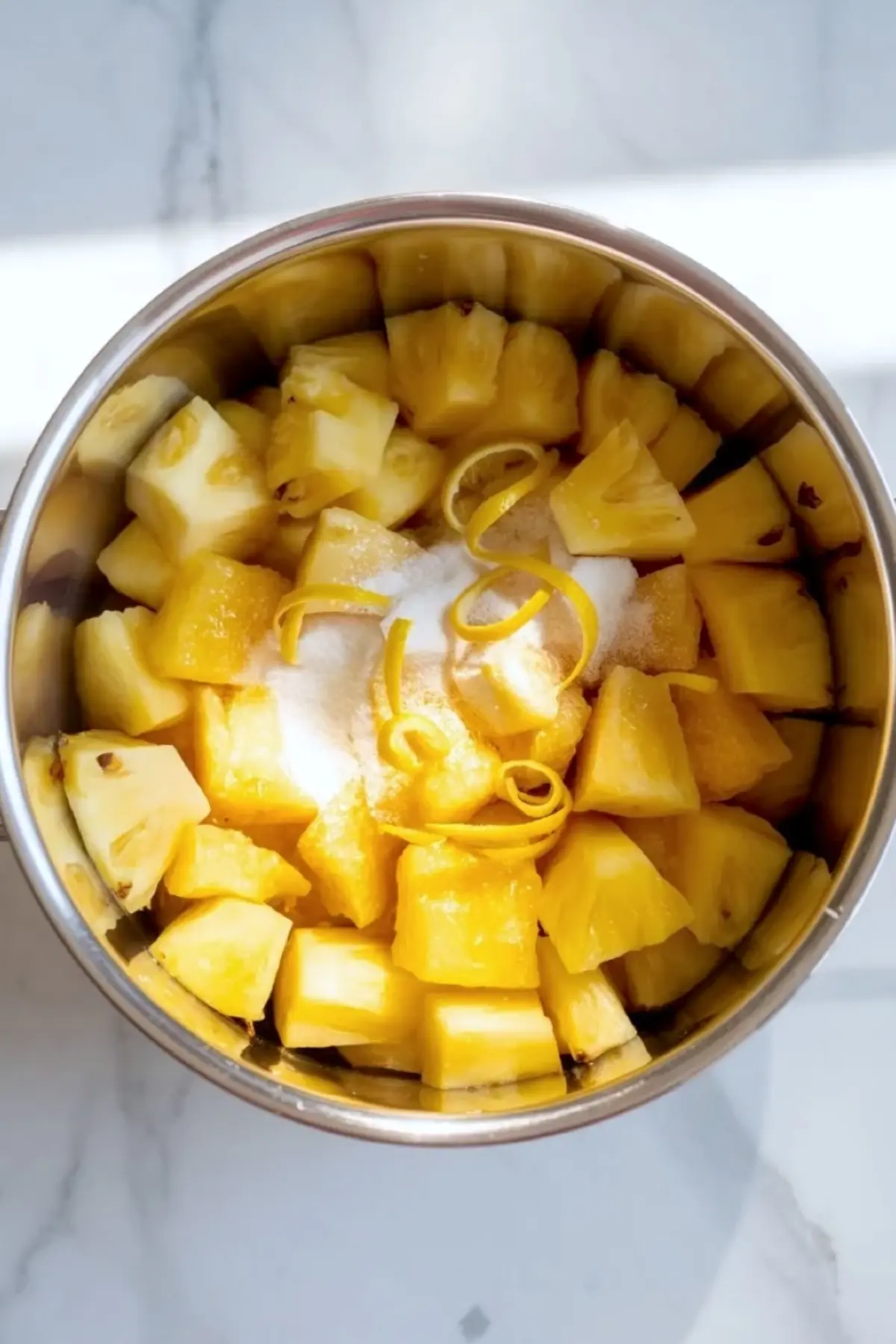 Pineapple chunks with sugar and lemon zest in a saucepan for homemade pineapple jam.