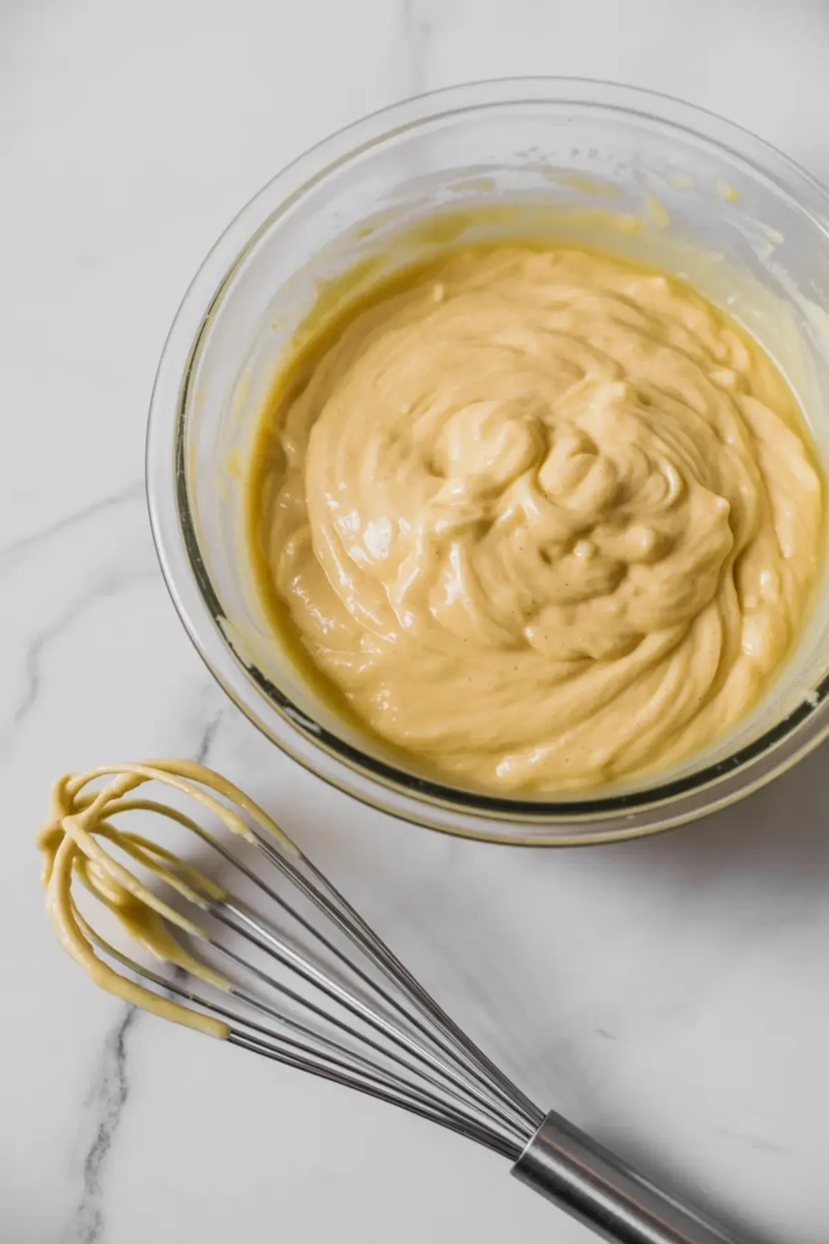 Glass bowl contains thick yellow cake batter beside a metal whisk on a marble surface. Smooth batter texture shows a step in homemade pineapple cake recipe preparation.
