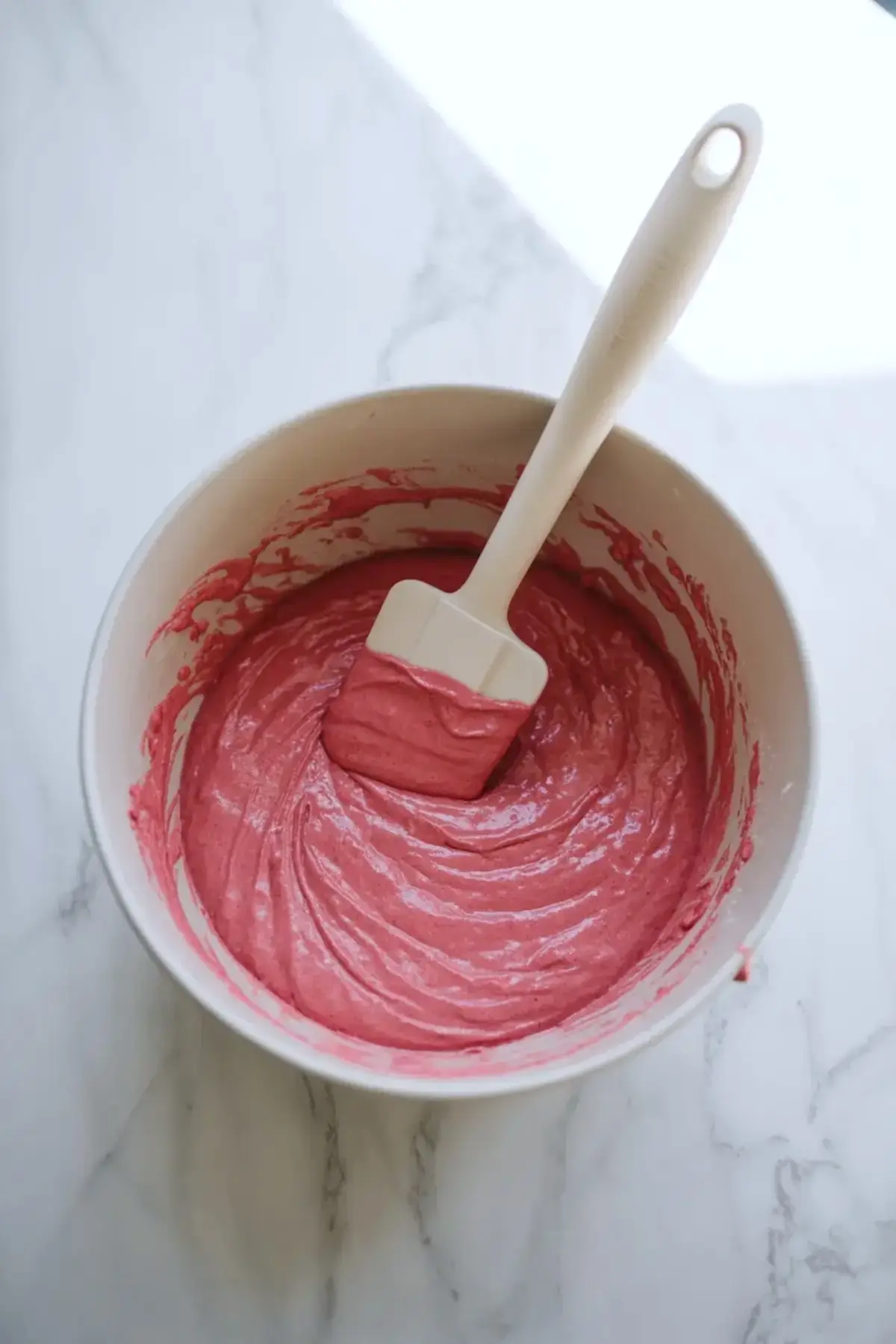 Spatula mixes smooth pink cake batter in a white bowl on a marble countertop, showing vibrant pink lemonade cake batter texture for a homemade cake recipe.
