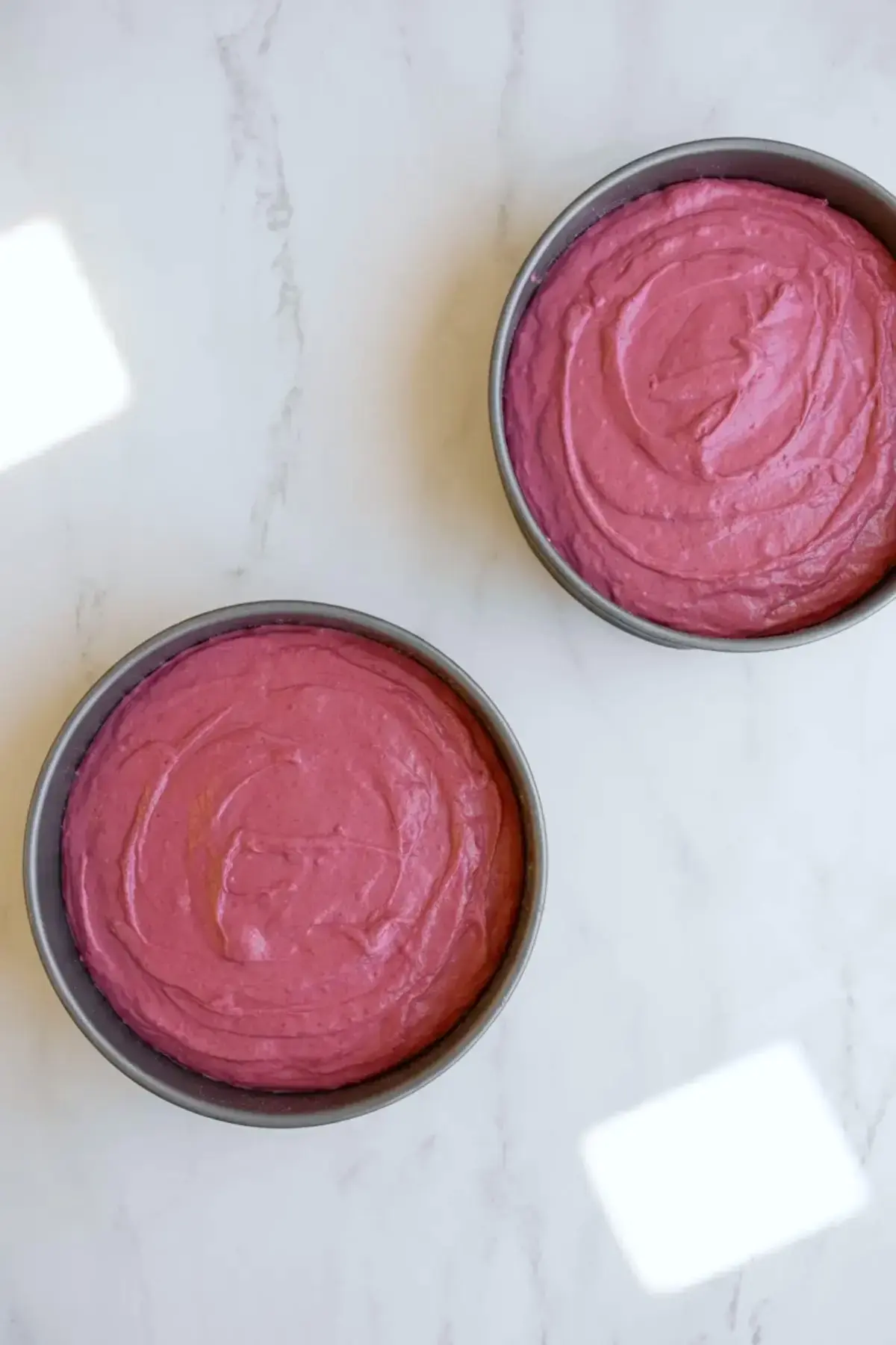Two round cake pans hold vibrant pink lemonade cake batter ready for baking on a white marble countertop during homemade layer cake preparation.
