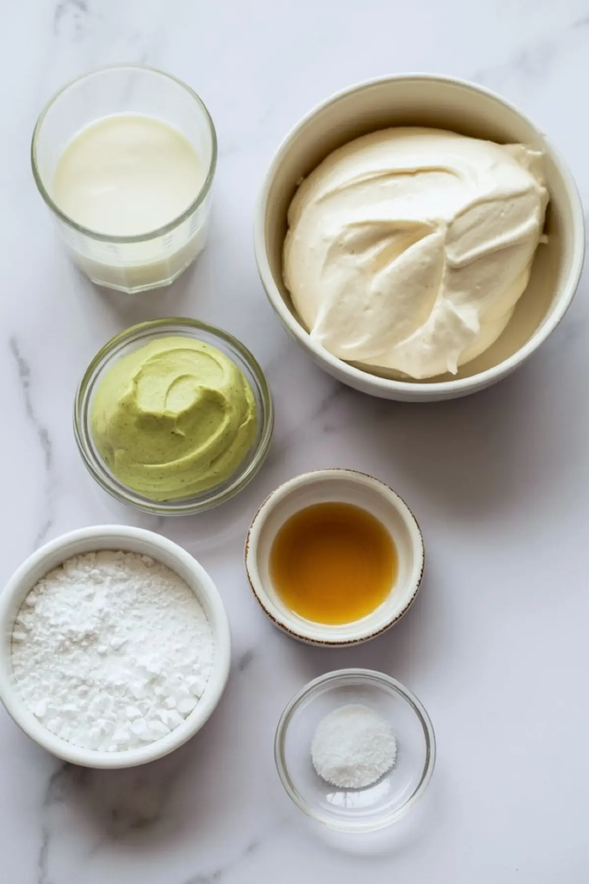 Measured ingredients for pistachio mascarpone frosting sit on a marble countertop, including mascarpone cheese, pistachio cream, powdered sugar, vanilla extract, milk, and salt in small bowls for an easy cake filling recipe.