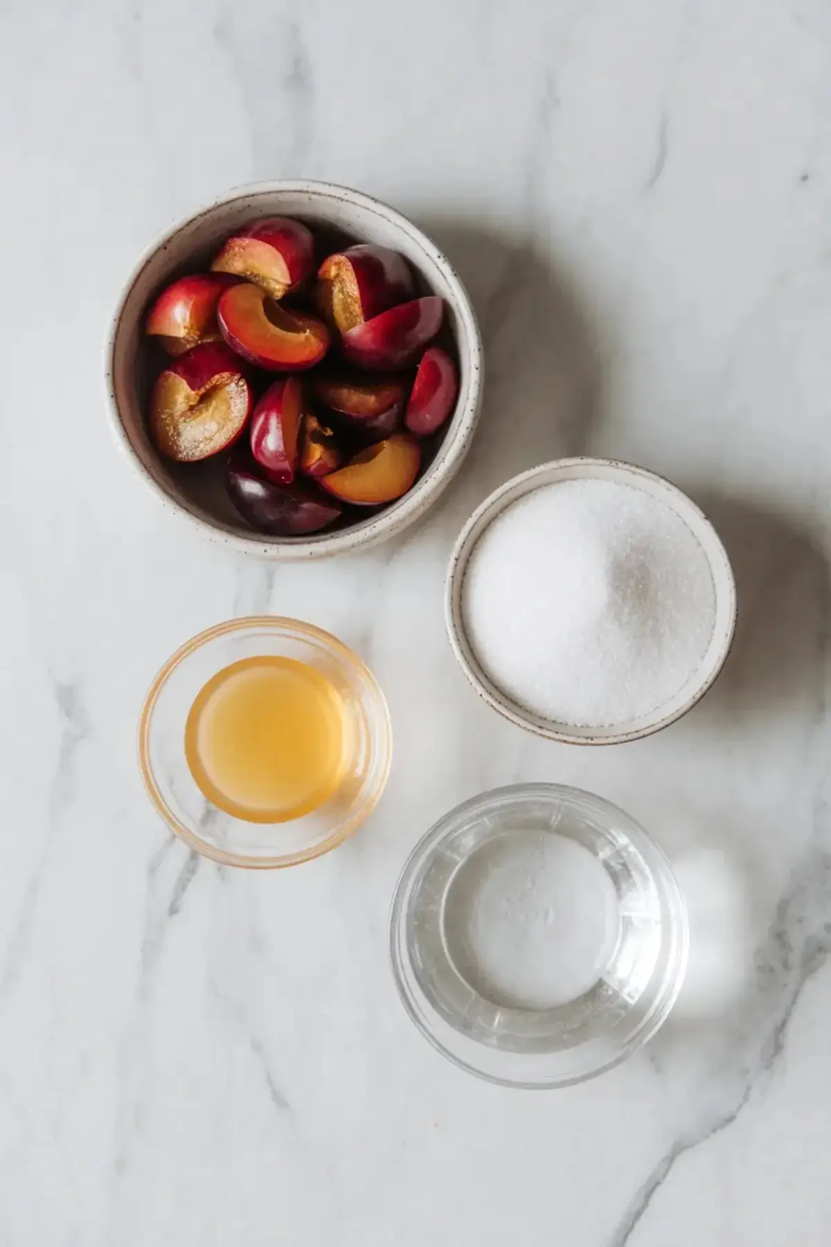 Plum jam ingredients sit on a marble surface with chopped plums, sugar, lemon juice, and water measured in small bowls.