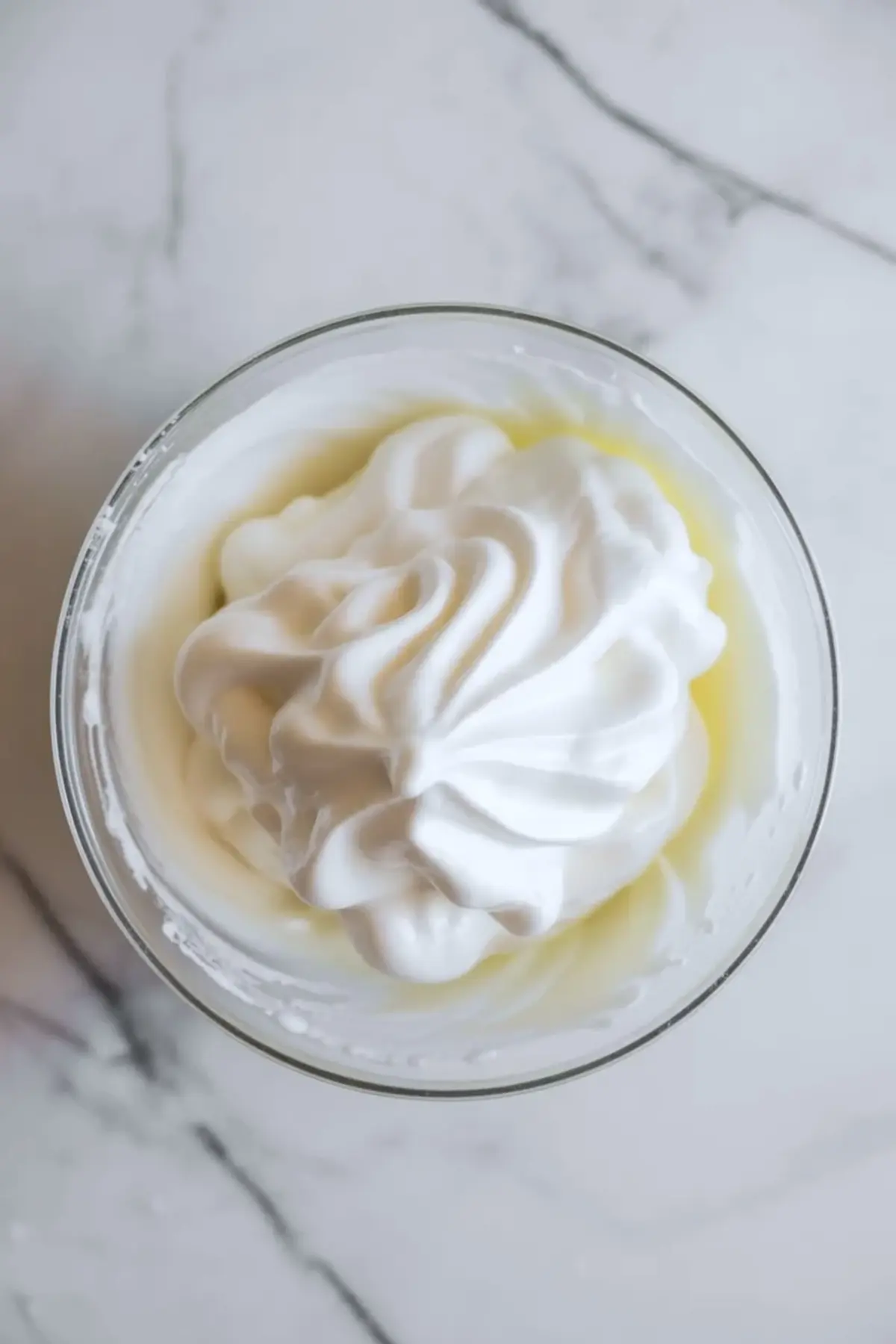 Whipped cream in a glass bowl on white marble, ready to be folded into raspberry mousse filling for cake.