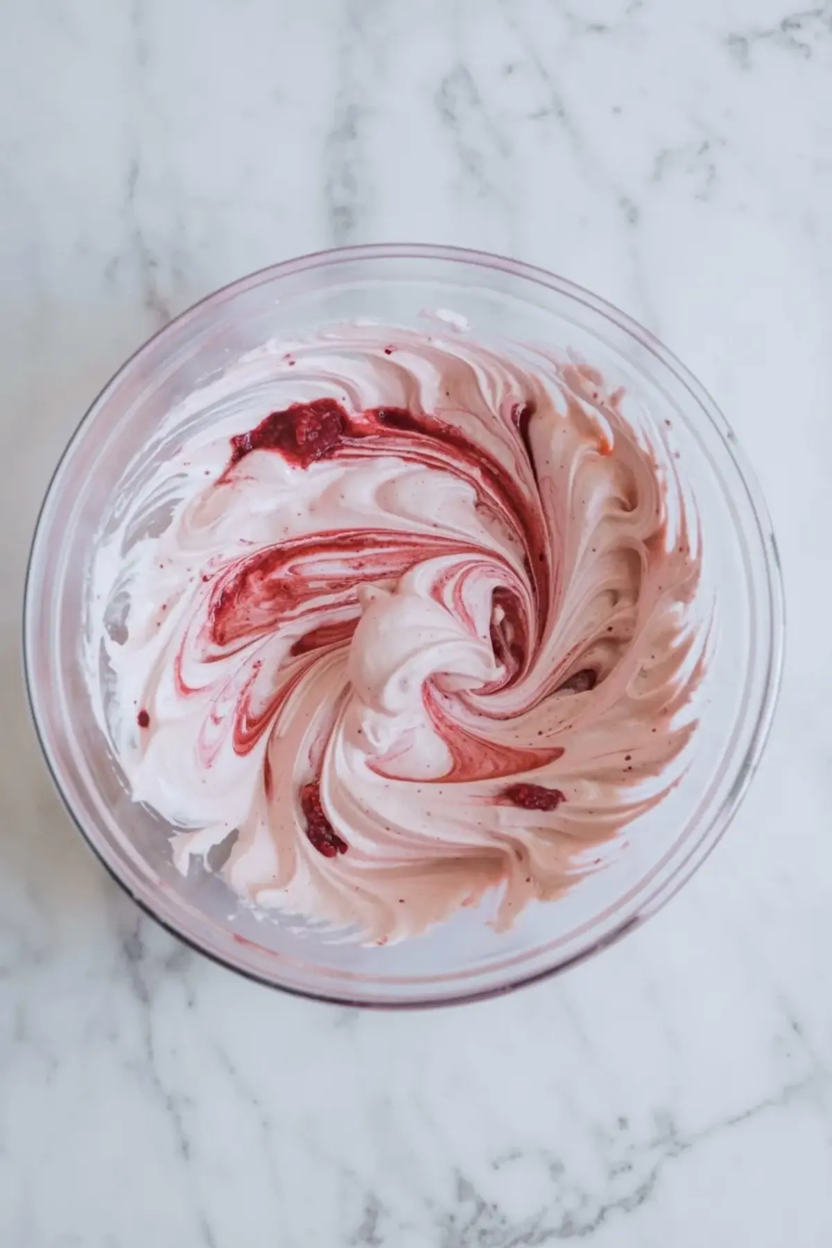 Raspberry puree folded into whipped cream in a glass bowl, creating swirls for raspberry mousse filling.