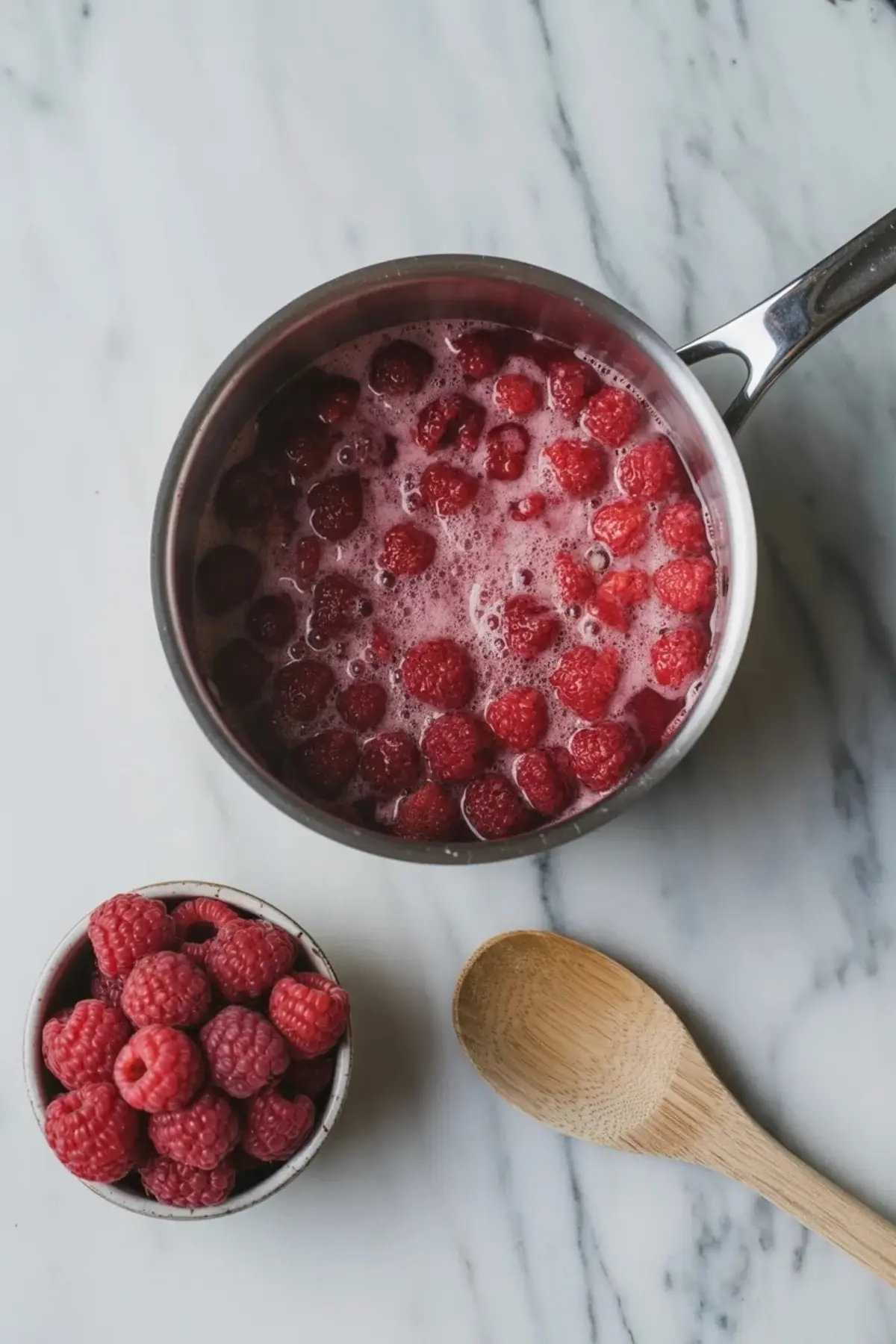 Fresh raspberries simmering in a saucepan on white marble for the fruit base of raspberry mousse filling.