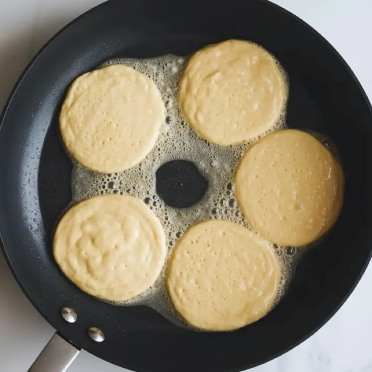 Five small pancakes cook in a buttered nonstick skillet with bubbling butter around the edges, showing golden sour cream pancake batter frying on the stovetop.
