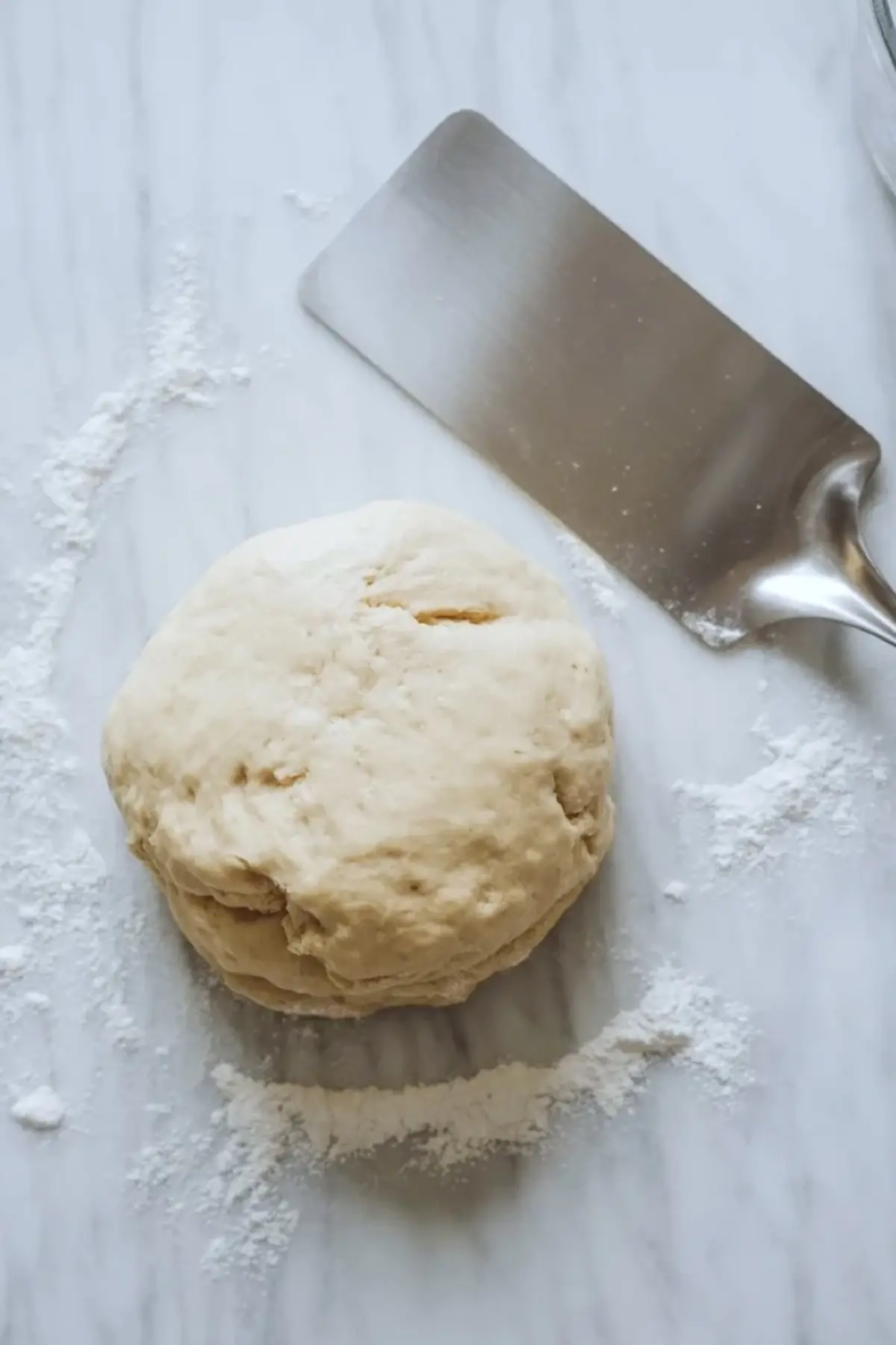 Round ball of scone dough resting on a floured marble surface with a metal bench scraper nearby, showing dough preparation for tender homemade scones.