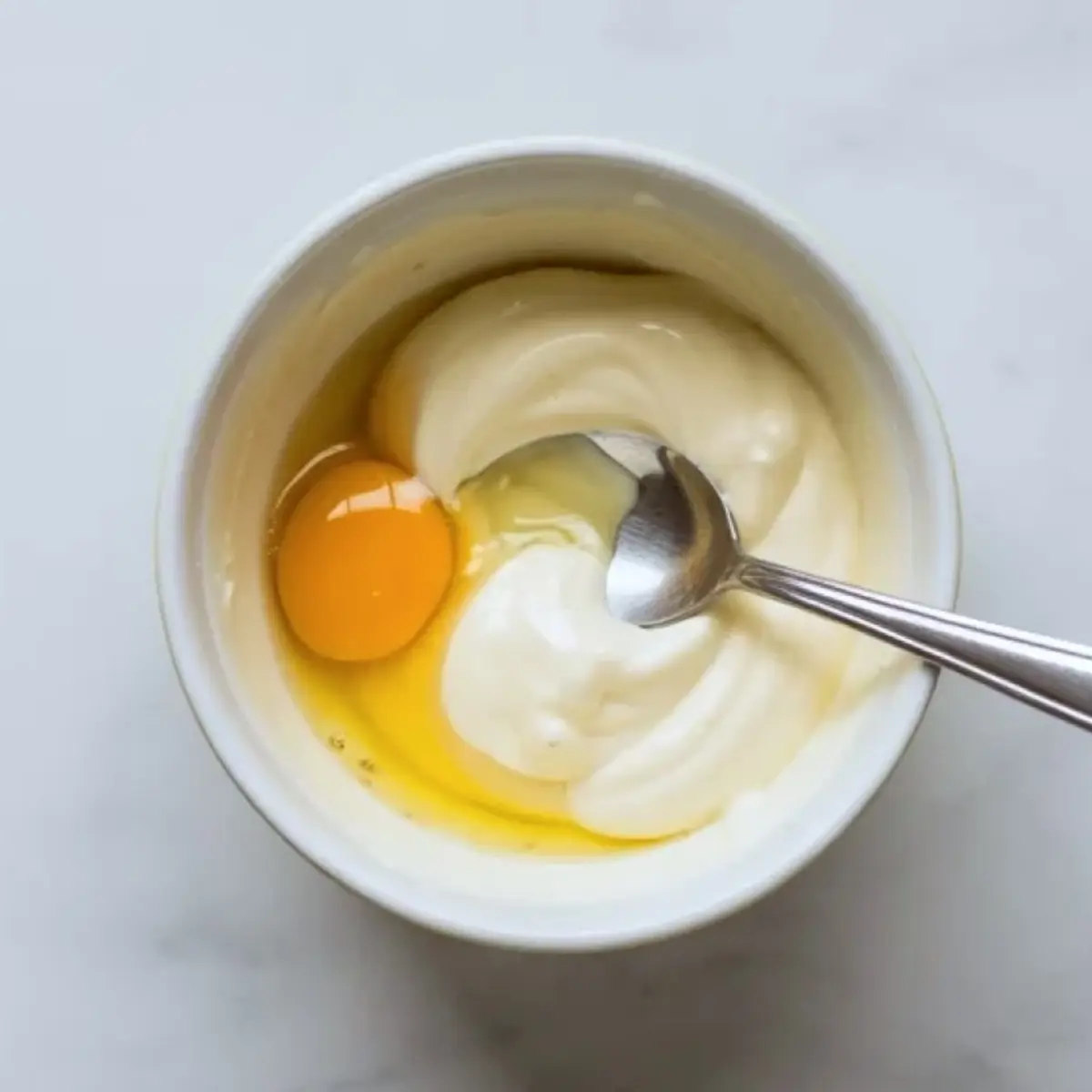 White bowl with sour cream and a whole egg being mixed with a spoon, showing wet ingredients for sour cream scones batter.