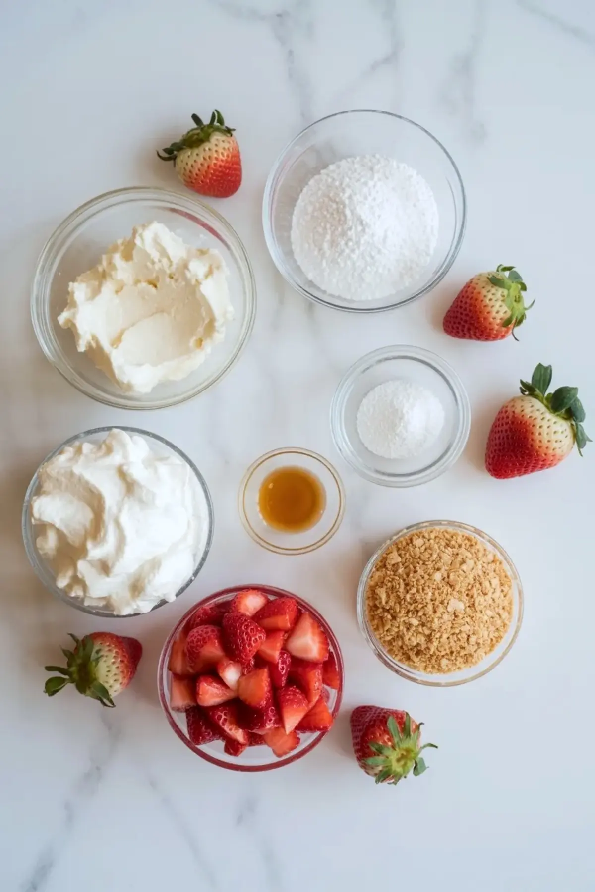 Ingredients for strawberry cheesecake fluff arranged on a white marble surface with cream cheese, whipped topping, fresh strawberries, sugar, vanilla, and graham cracker crumbs.