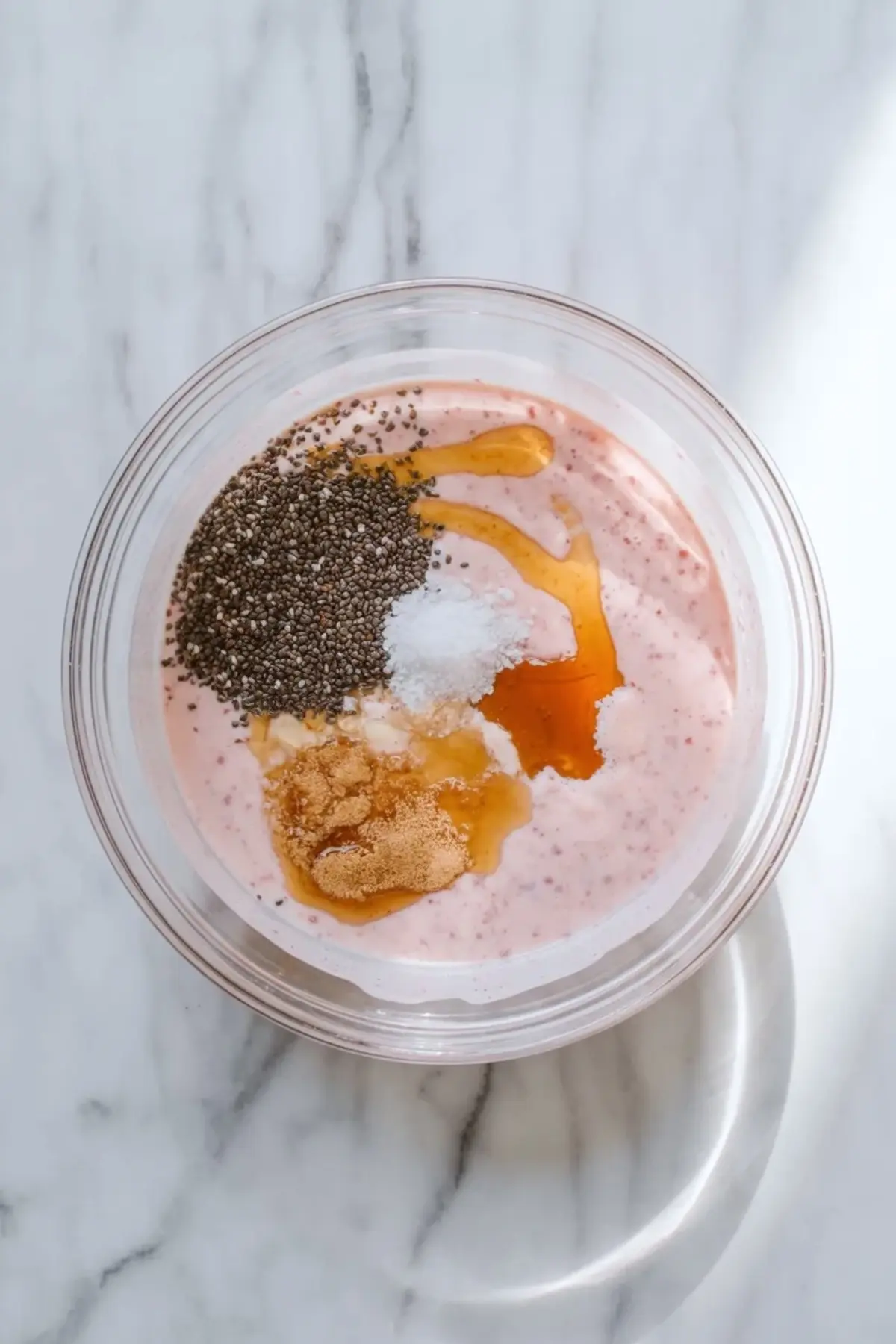 Top view of a bowl with pink strawberry milk mixture topped with chia seeds, honey drizzle, vanilla extract, and sugar on a marble countertop for chia pudding preparation.