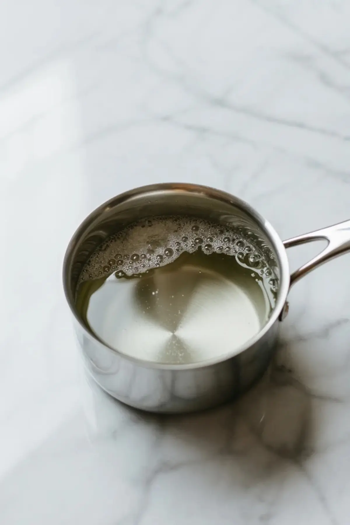 Simple syrup warming in a small saucepan for strawberry lemon mint sorbet on a white marble surface.