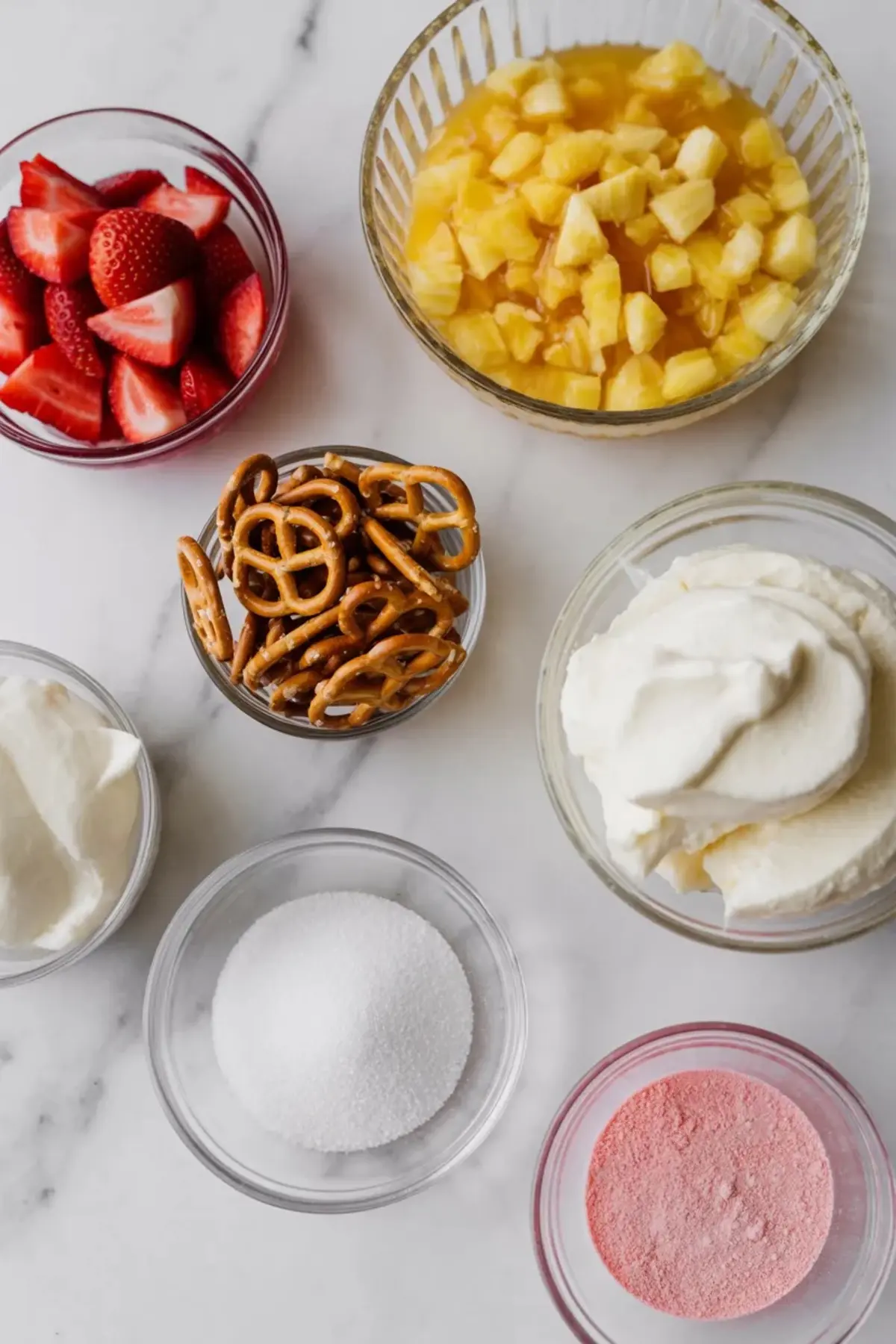 Ingredients for strawberry pretzel fluff arranged on a white marble surface with strawberries, crushed pineapple, pretzels, whipped topping, cream cheese, sugar, and strawberry gelatin mix.