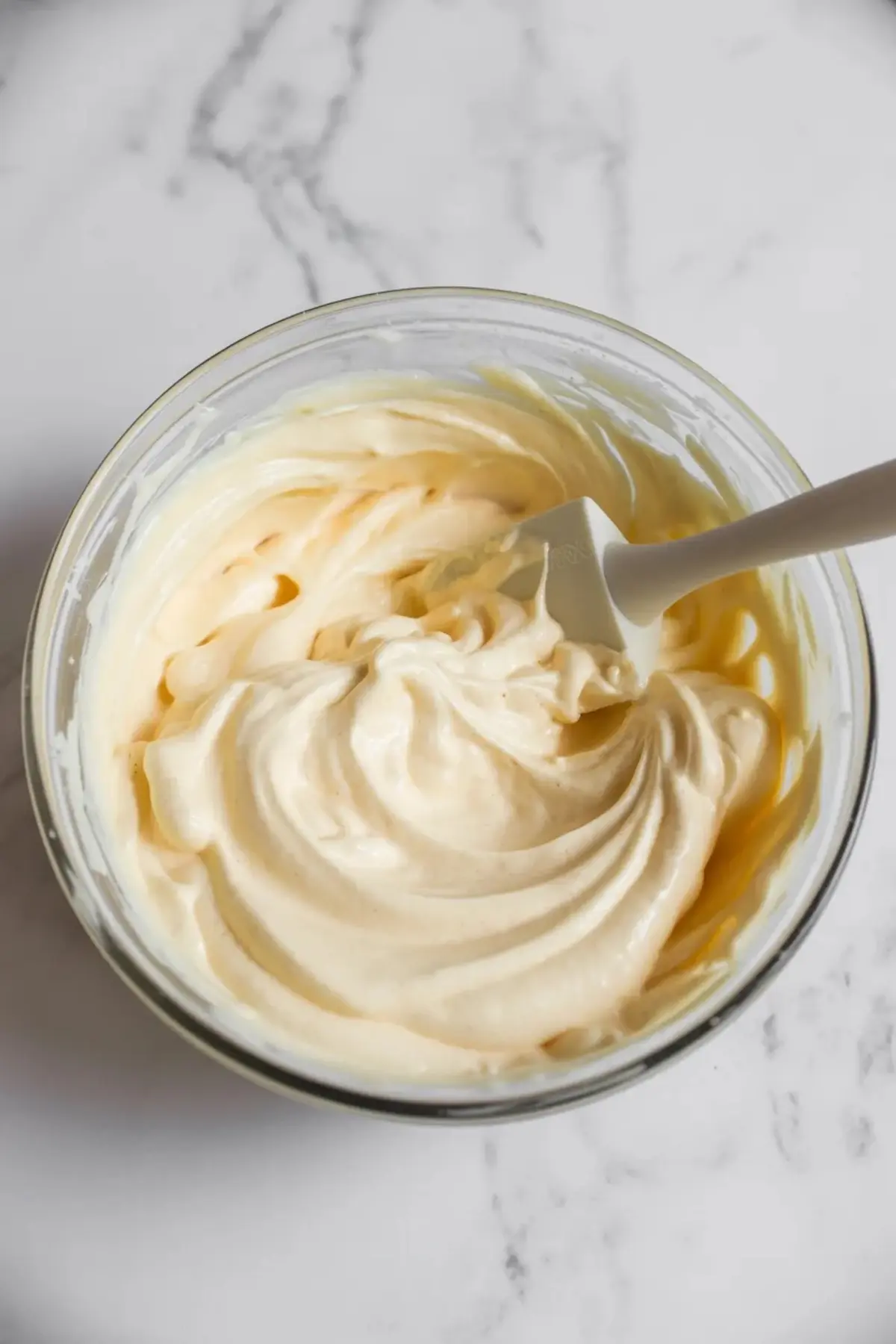 Vanilla mousse cake filling swirled in a glass bowl with a spatula, showing the creamy texture on a white marble surface.