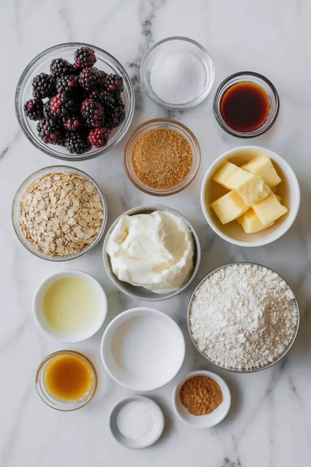 Ingredients for blackberry crisp with whipped mascarpone arranged on marble with fresh blackberries, oats, flour, butter, sugars, mascarpone, cream, lemon juice, vanilla, cinnamon, and salt.