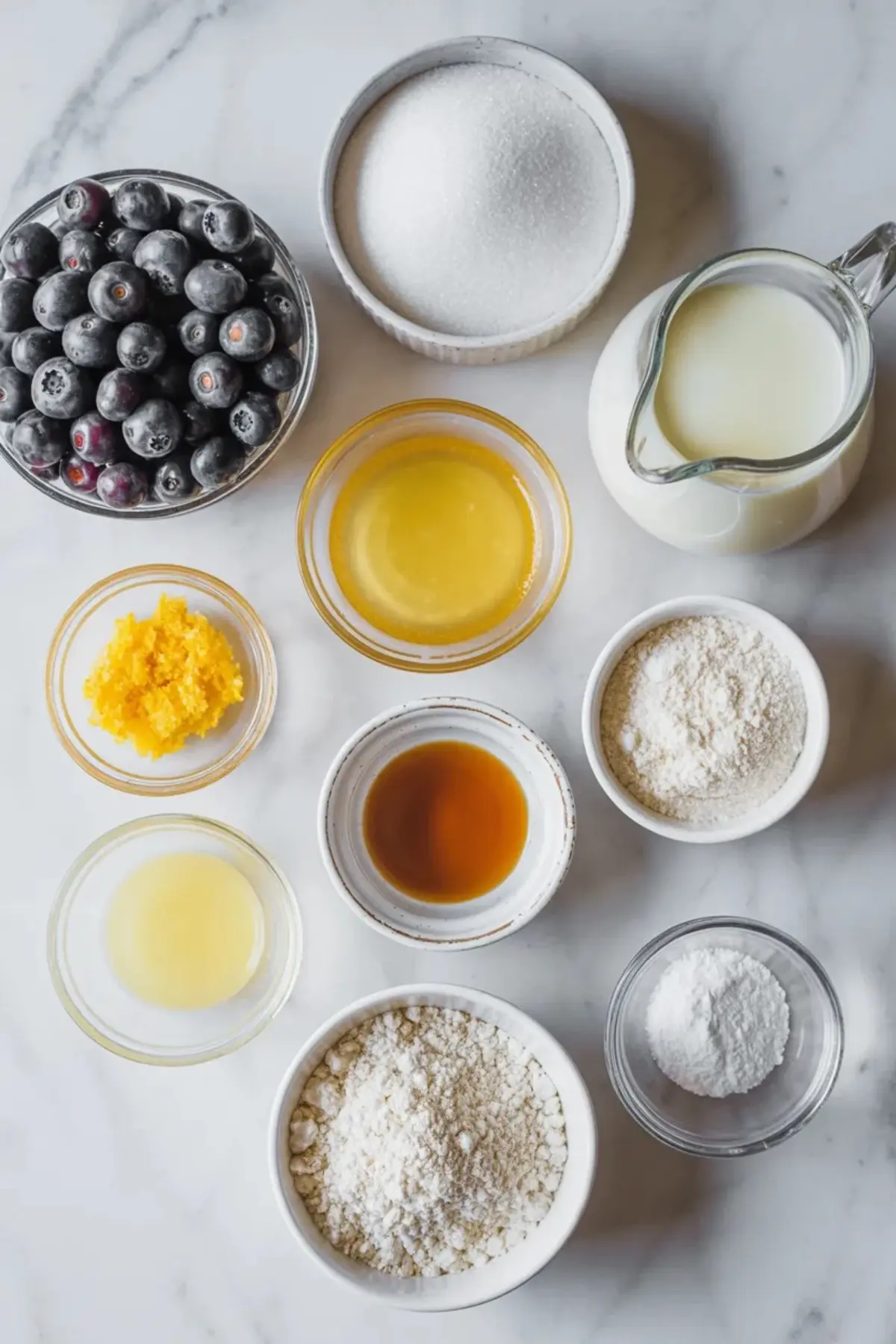 Ingredients for blueberry cobbler arranged on marble with blueberries, flour, sugar, butter, milk, baking powder, and vanilla.