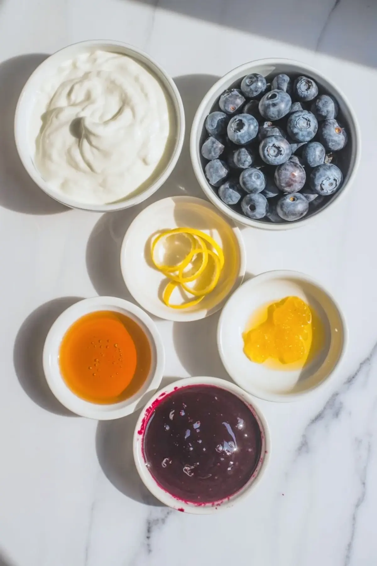 Blueberry lemon yogurt bark ingredients arranged in bowls with yogurt, blueberries, lemon zest, honey, lemon curd, and blueberry sauce.