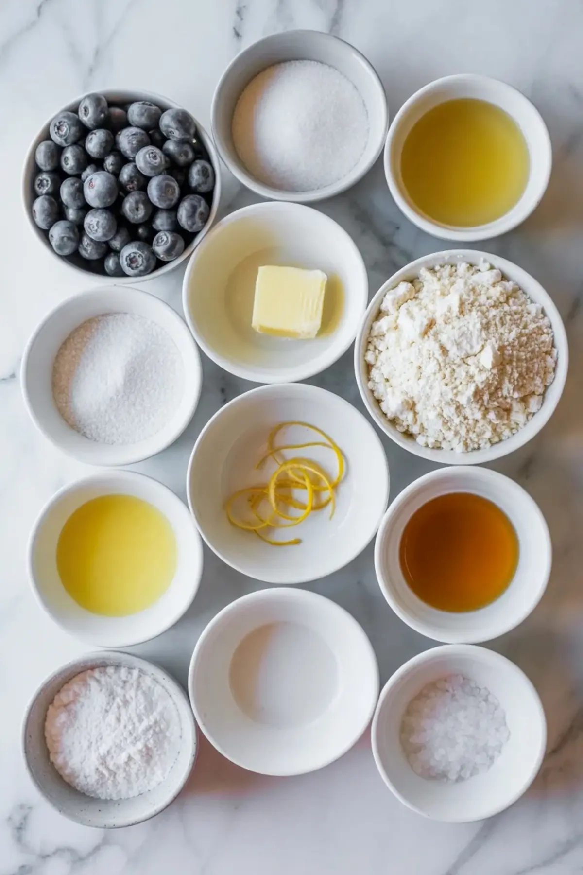 Ingredients for blueberry pie bars arranged on white marble with blueberries, flour, sugar, butter, lemon juice, lemon zest, vanilla, powdered sugar, and salt.