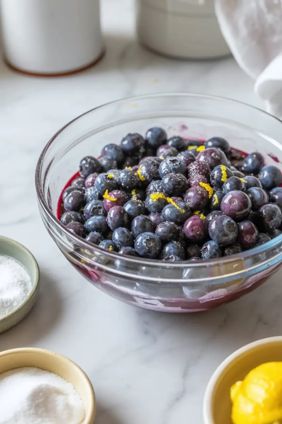 Fresh blueberry filling for blueberry pie bars in a glass bowl with lemon zest and juices on a marble surface.