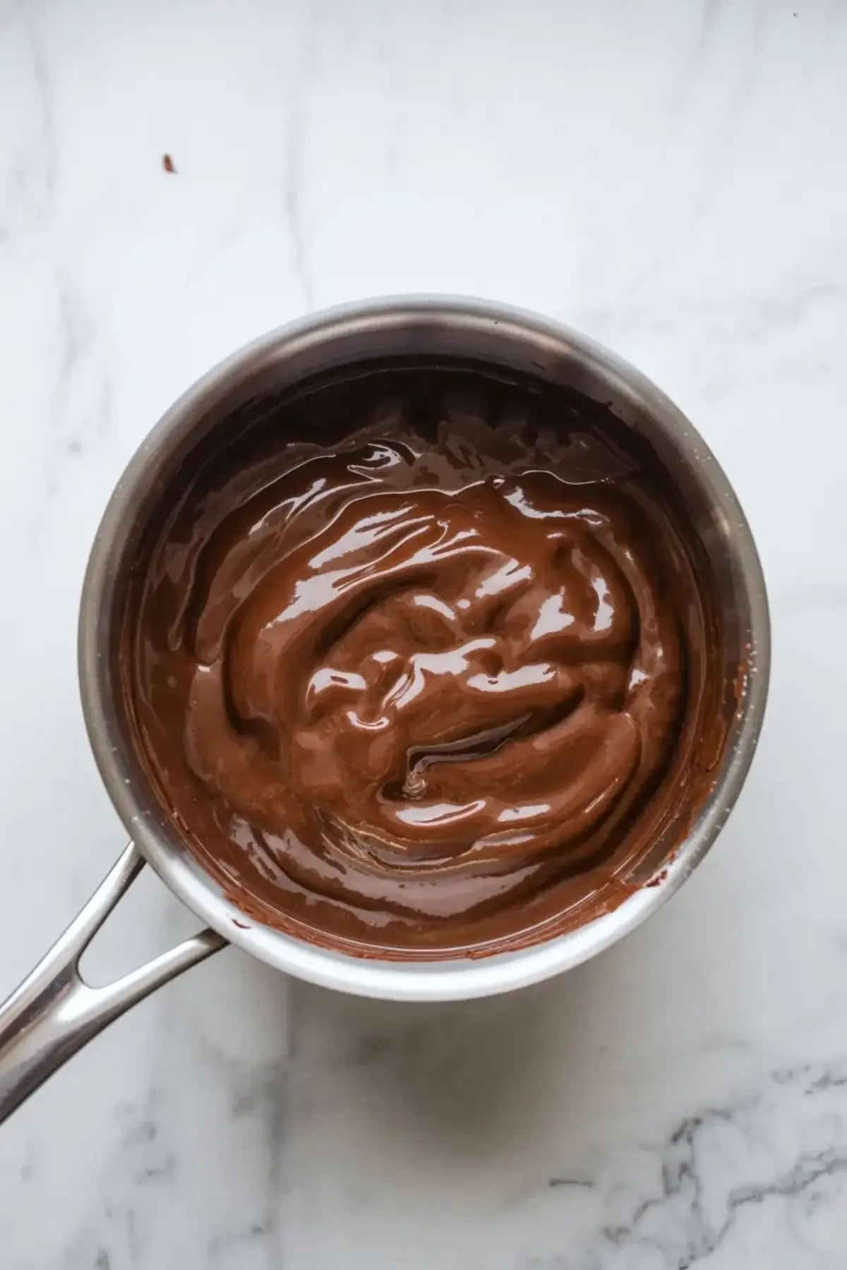 Smooth chocolate filling for chocolate vanilla cream pie bars in a metal saucepan on a marble surface.