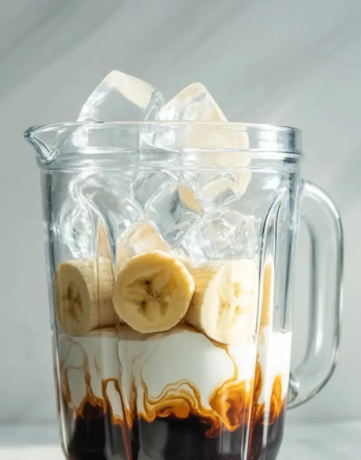 Blender jar filled with coffee, coconut milk, banana slices, and ice before blending the coconut coffee smoothie.