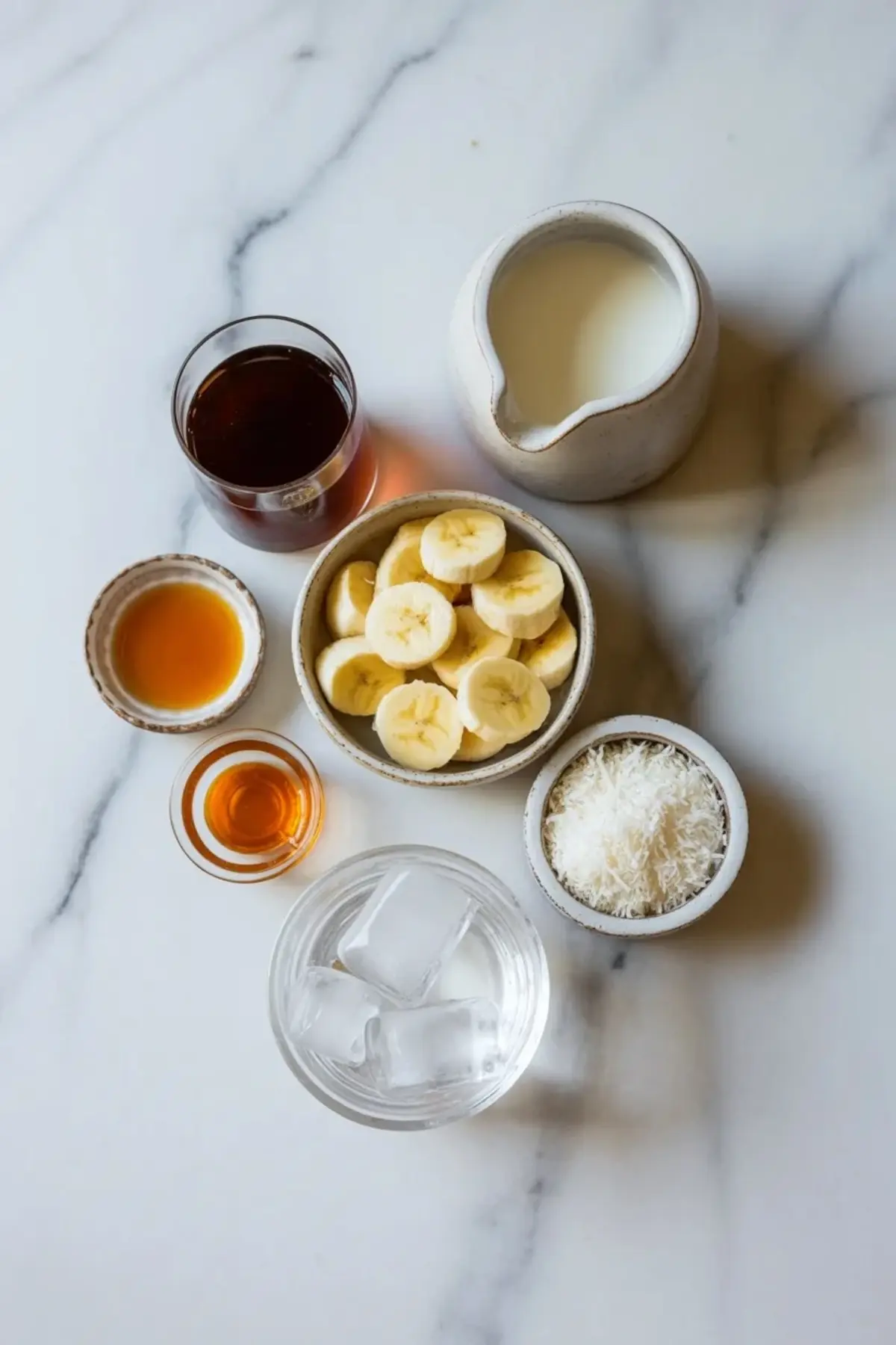 Ingredients for coconut coffee smoothie arranged on marble with brewed coffee, coconut milk, banana slices, shredded coconut, sweetener, and ice.