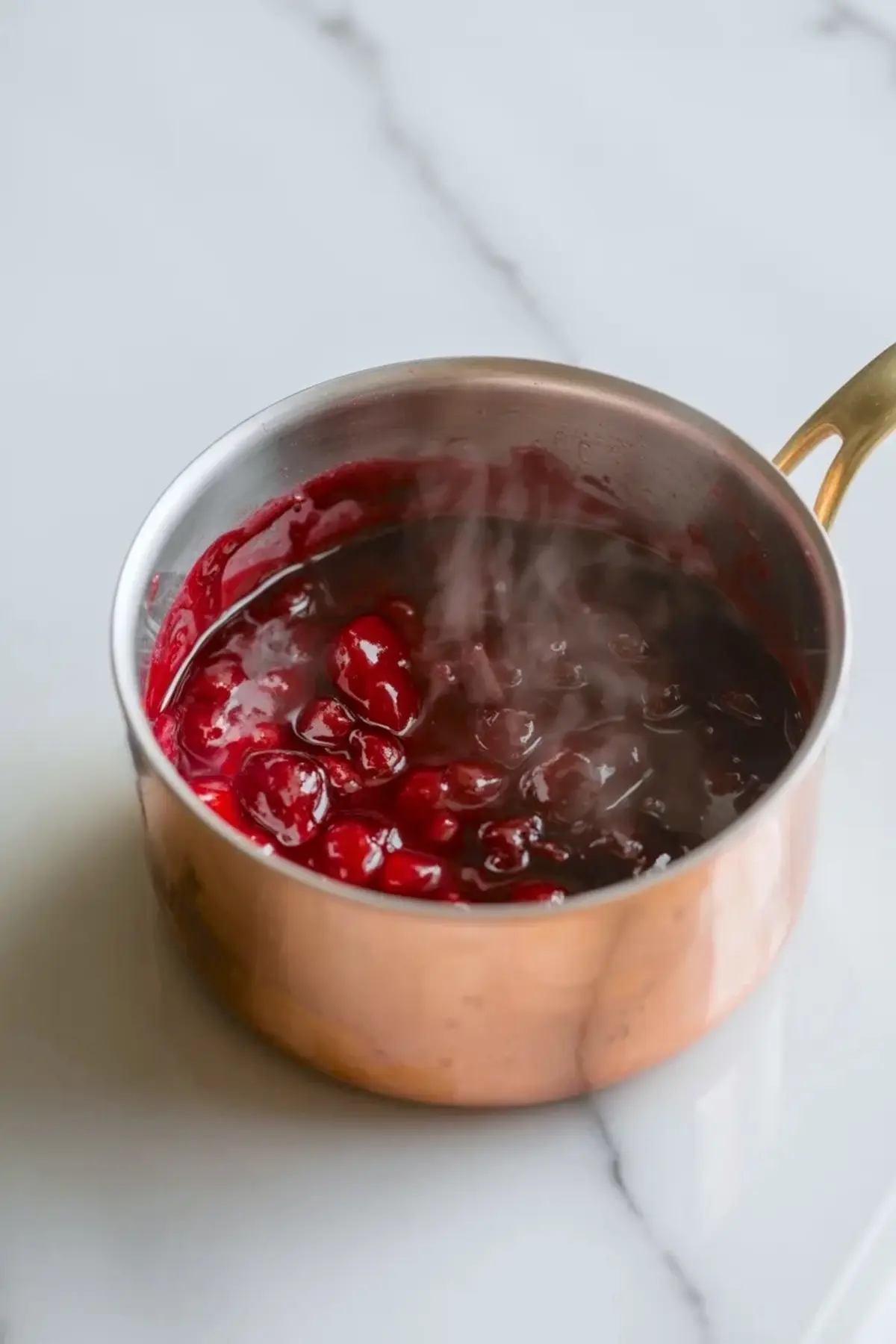 Cherry sauce simmering in a small copper pan for topping coconut ice cream on a white marble surface.