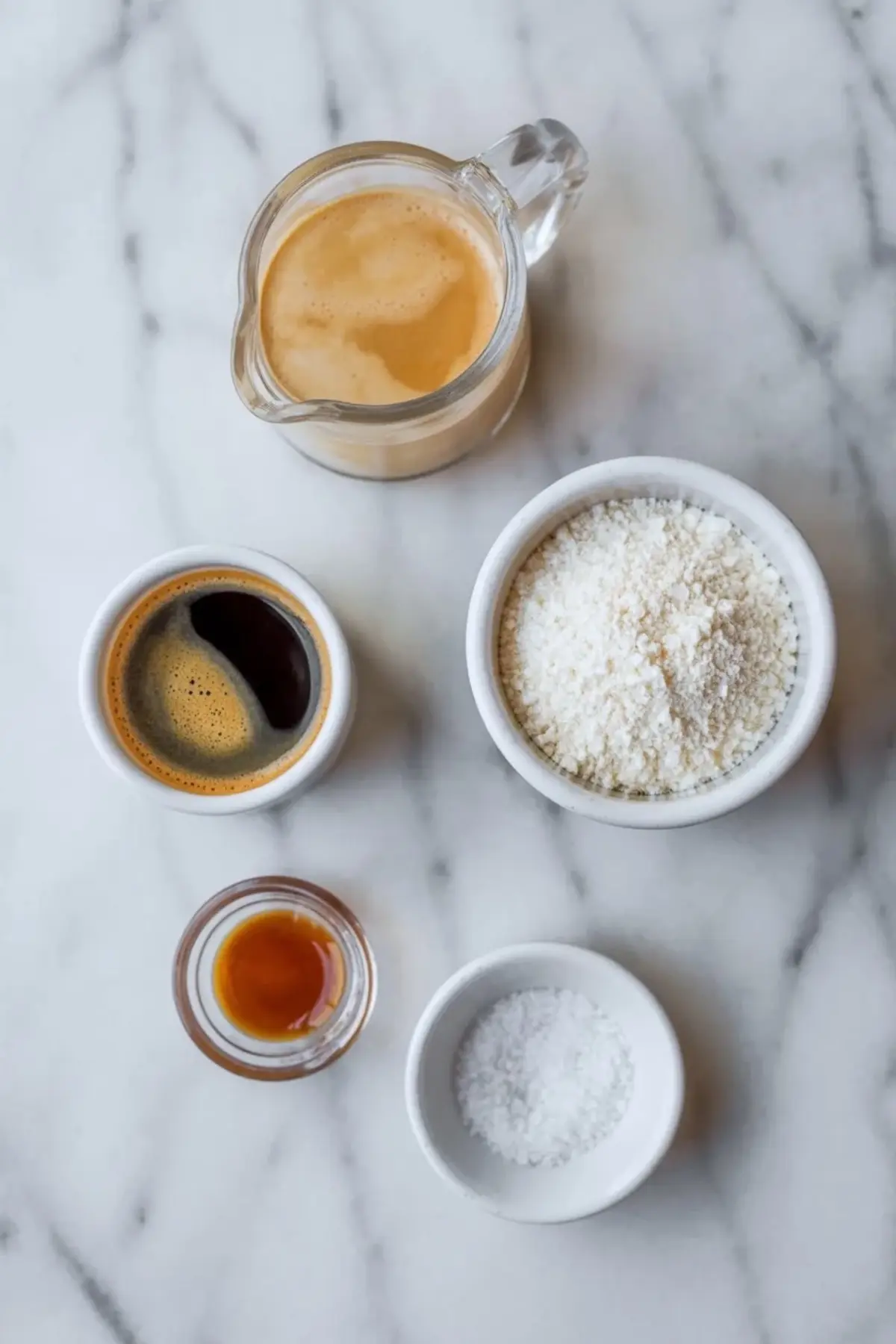 Ingredients for espresso whipped cream arranged on white marble with heavy cream, espresso, powdered sugar, vanilla, and salt.
