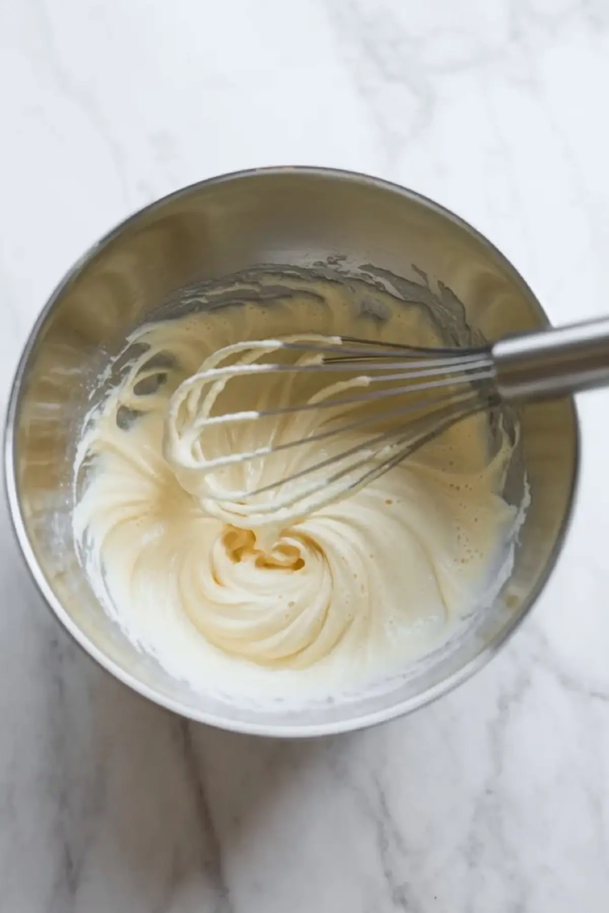 Espresso whipped cream being whisked in a metal bowl until it starts to thicken on a white marble surface.