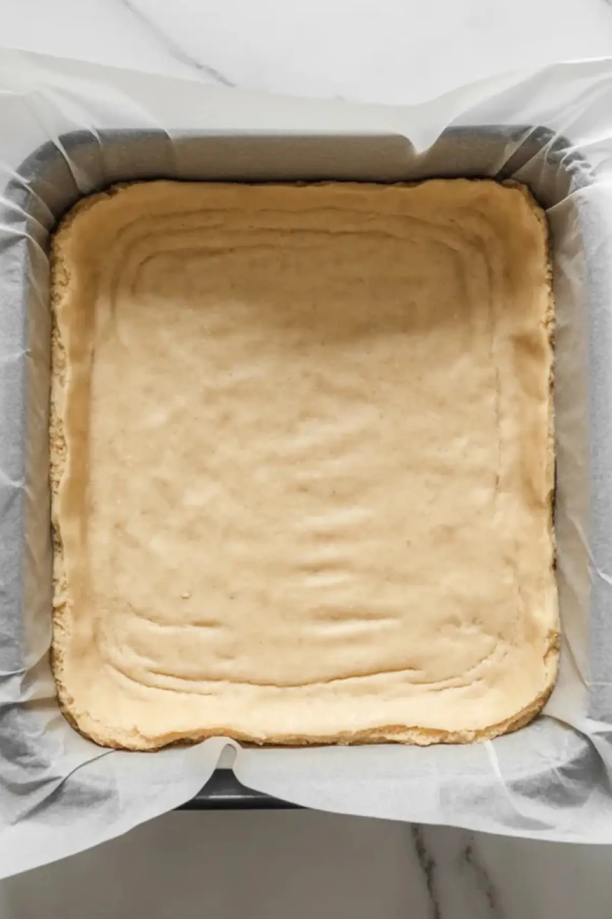 Shortbread crust pressed into a parchment lined square pan for lemon bars on a white marble surface.