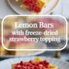 Lemon bars shown in a 2 to 3 hero image with several bars above and one plated bar below topped with freeze-dried strawberries.