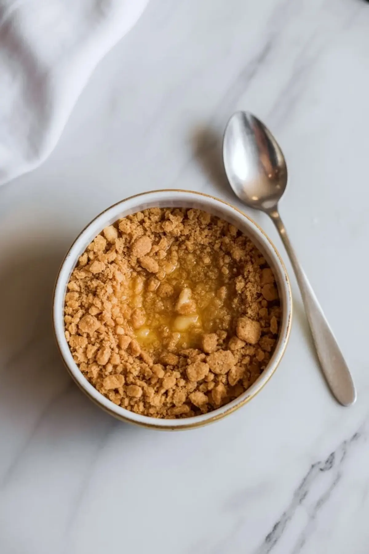 An individual lemon cheesecake cup sits in a small ramekin with graham crumbs visible on top beside a spoon on white marble.
