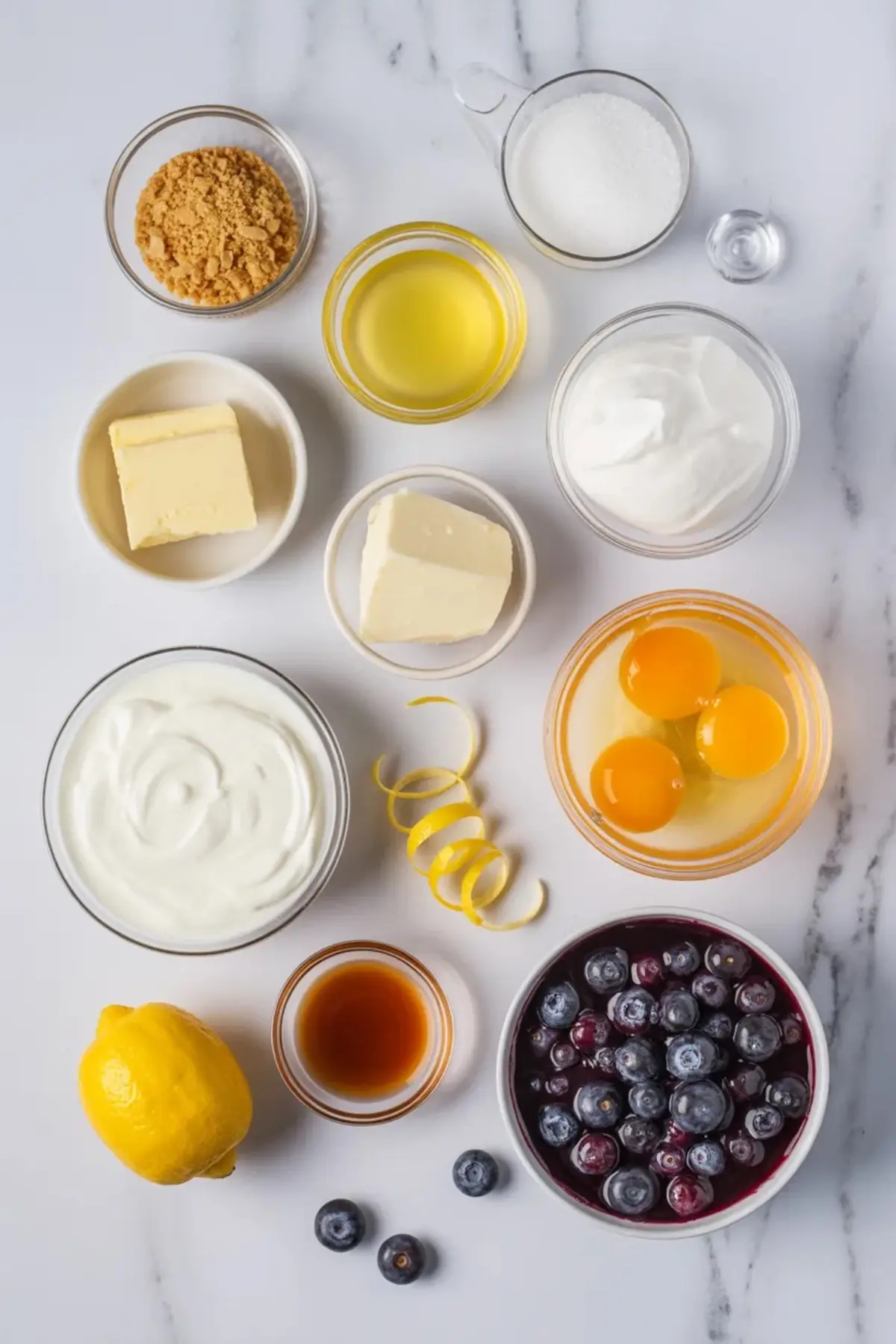 Ingredients for lemon blueberry cheesecake cups are arranged on white marble with cream cheese, sour cream, eggs, lemon, vanilla, graham crumbs, sugar, and blueberries.