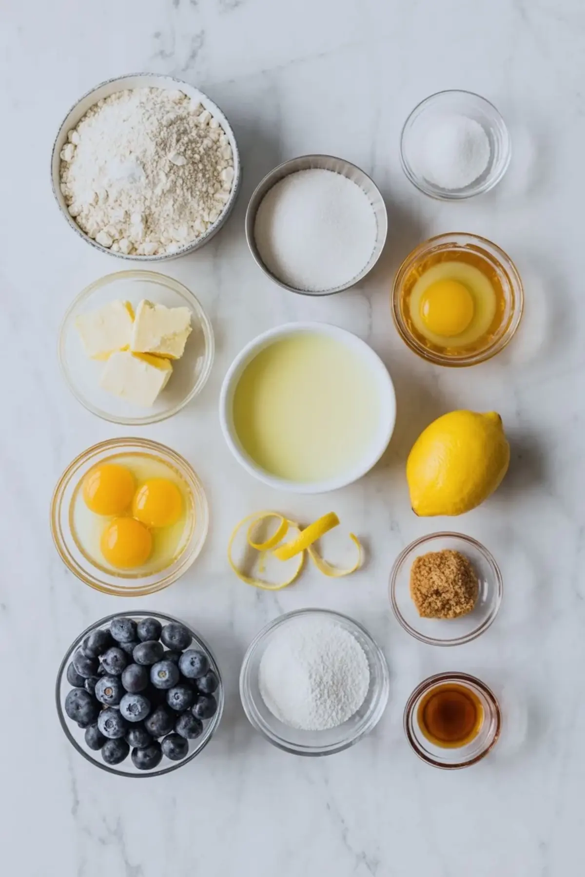 Ingredients for lemon blueberry crumb bars arranged on white marble with flour, sugar, butter, eggs, lemon, blueberries, vanilla, and brown sugar.