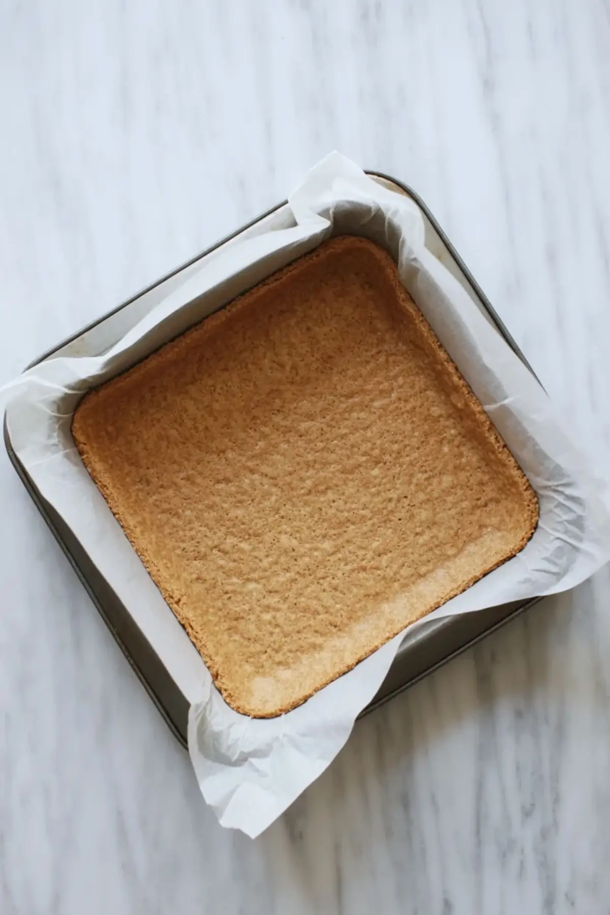 A baked graham cracker crust for orange creamsicle bars sits in a parchment lined square pan ready for the creamy filling.