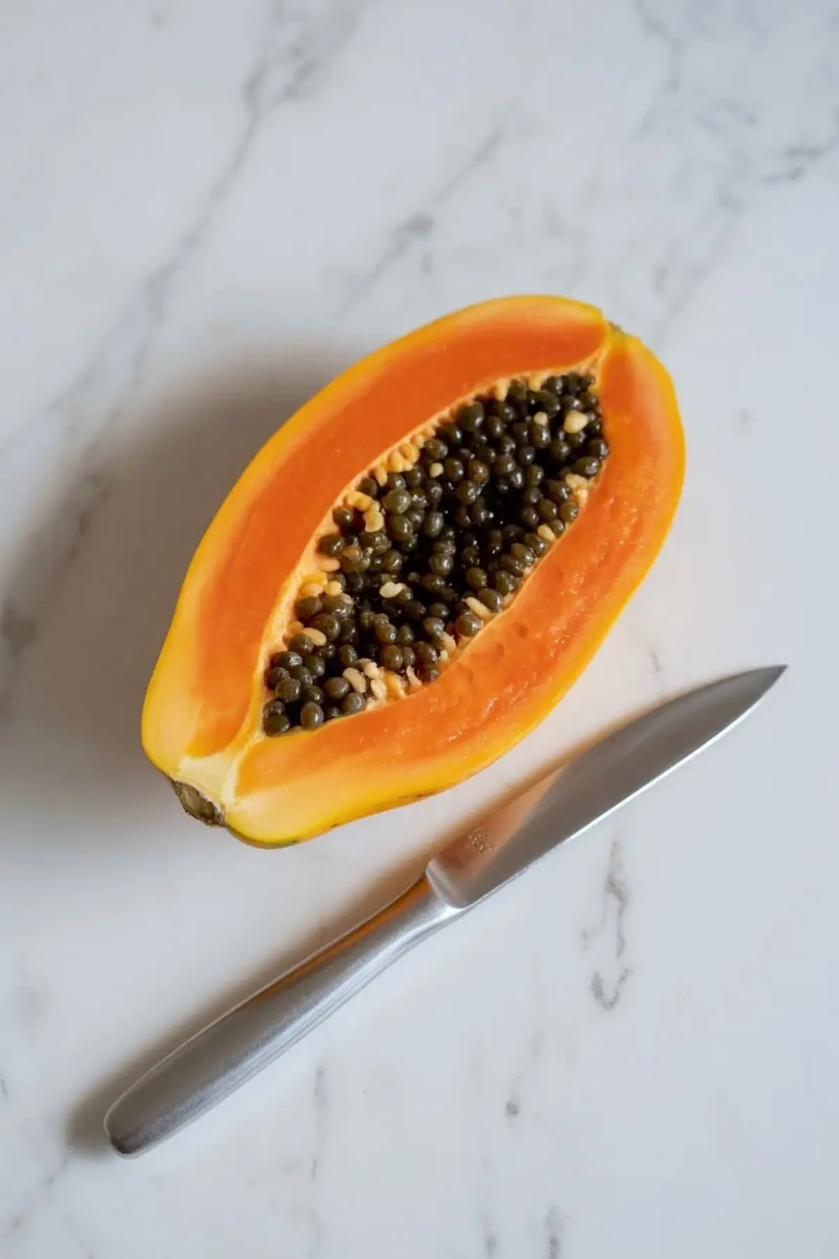 Ripe papaya cut in half with seeds still inside beside a knife on white marble for papaya lime boats preparation.