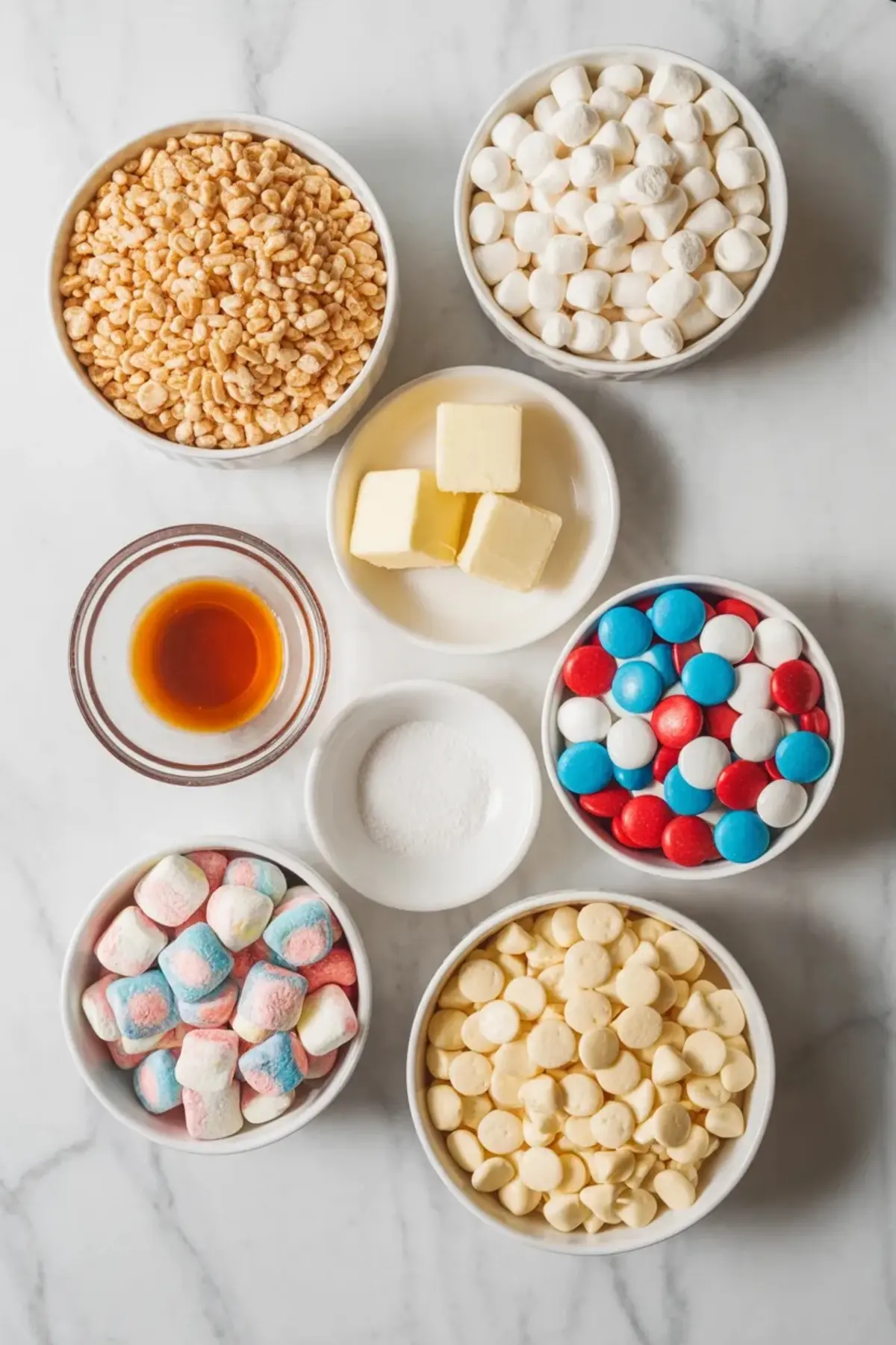 Ingredients for patriotic Rice Krispie treats including cereal, marshmallows, butter, vanilla, white chocolate chips, and red, white, and blue candy pieces.
