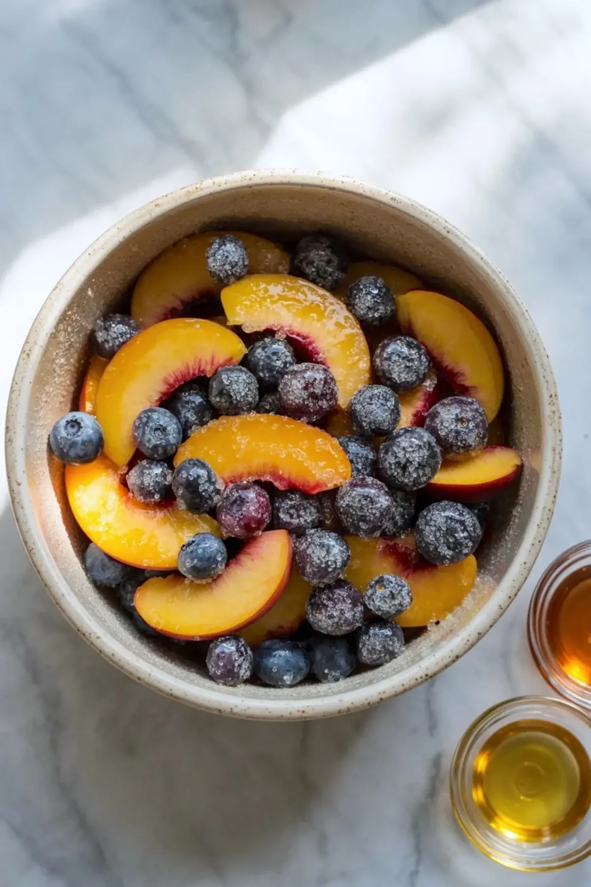 Peaches and blueberries for peach blueberry galette tossed in a bowl with sugar while small bowls of honey and vanilla sit nearby.