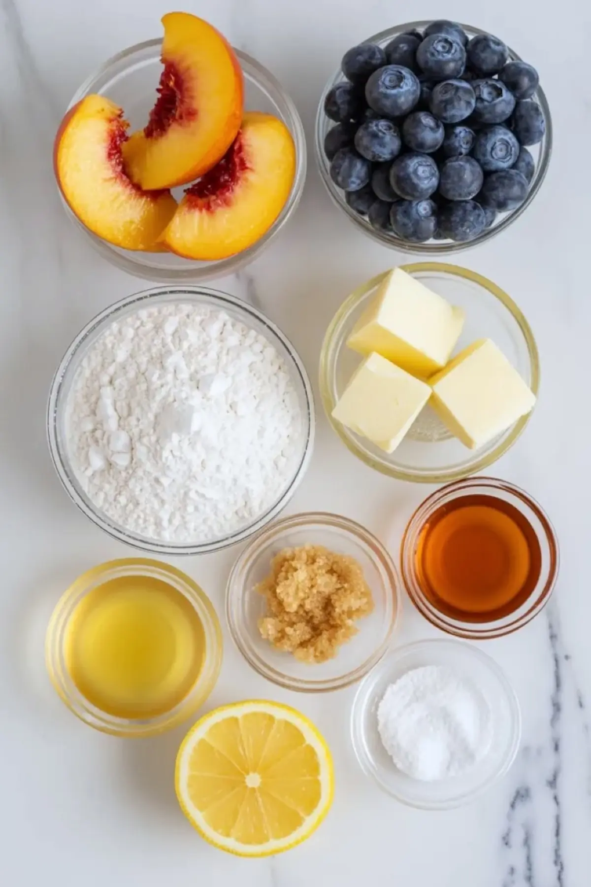 Ingredients for peach blueberry galette arranged on white marble with sliced peaches, fresh blueberries, flour, butter, lemon, sugar, vanilla, and cornstarch.